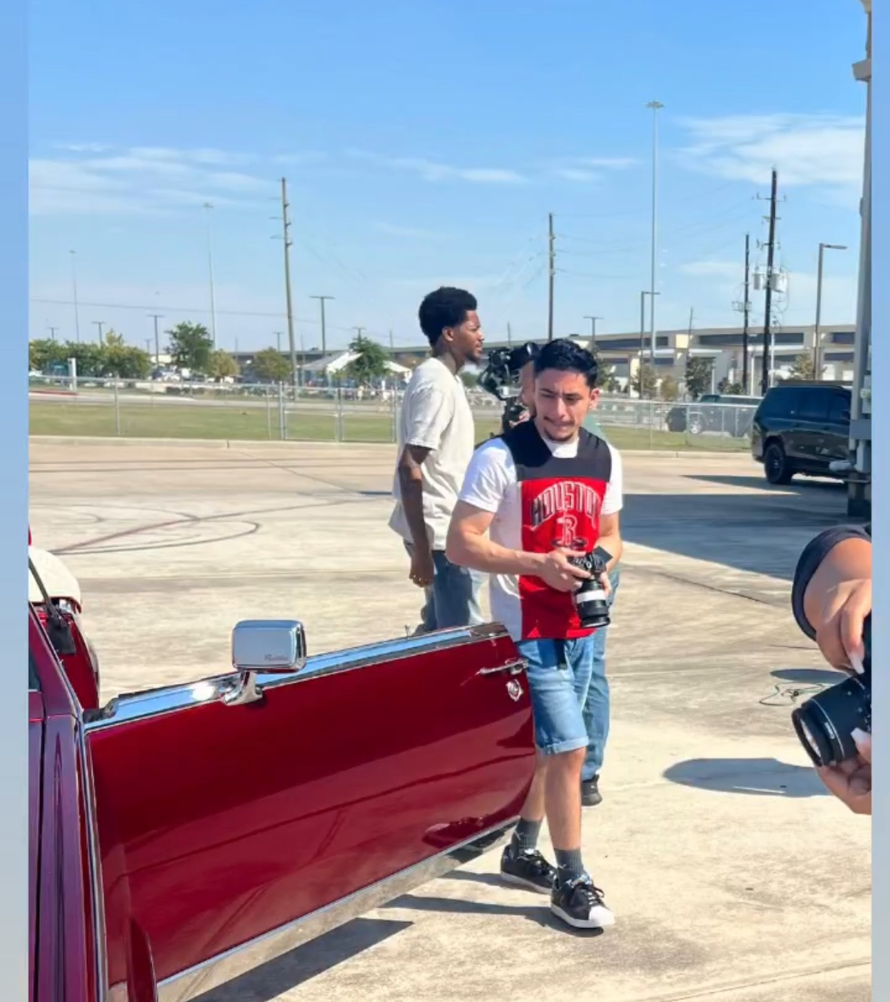 Two boys standing outdoors near a red vehicle, with a parking lot and blue sky in the background.