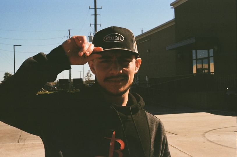 Person wearing a cap and hoodie, smiling outdoors with buildings and power lines in background.