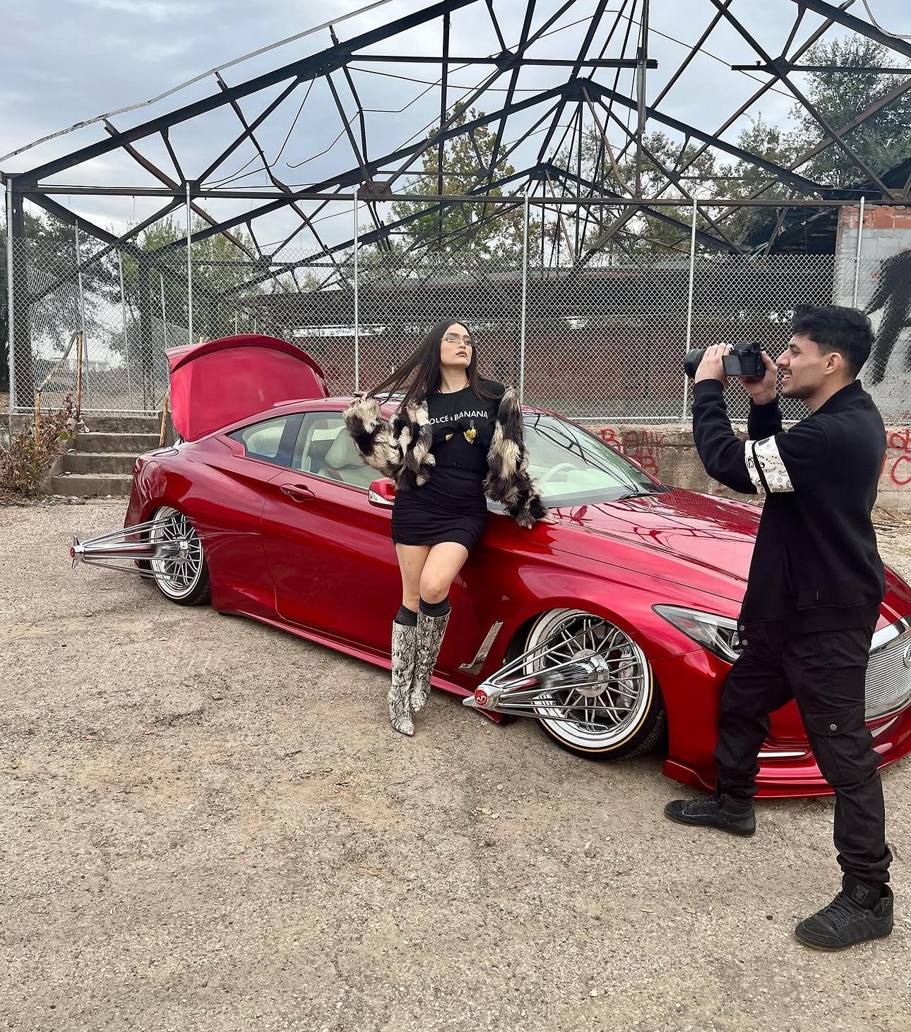 Person taking a photo of a woman posing next to a red sports car with an open trunk, outdoors on a dirt surface.
