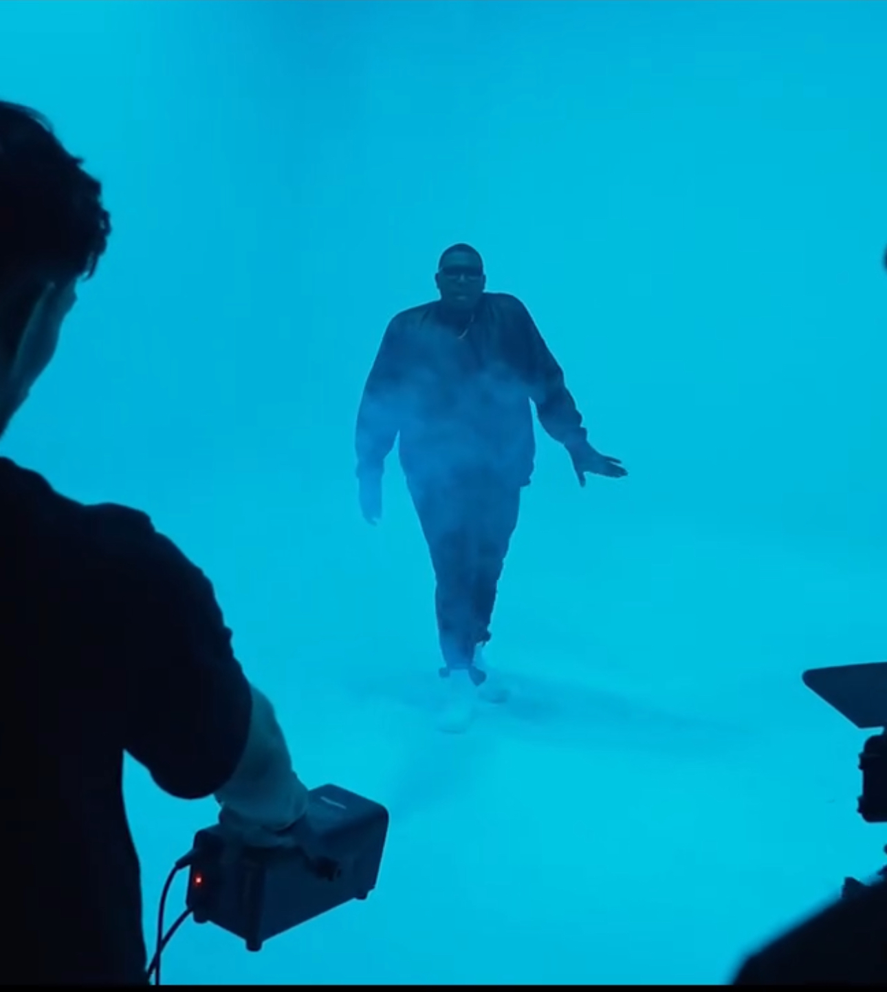 Person walking towards camera in foggy blue environment, with two people filming from sides.