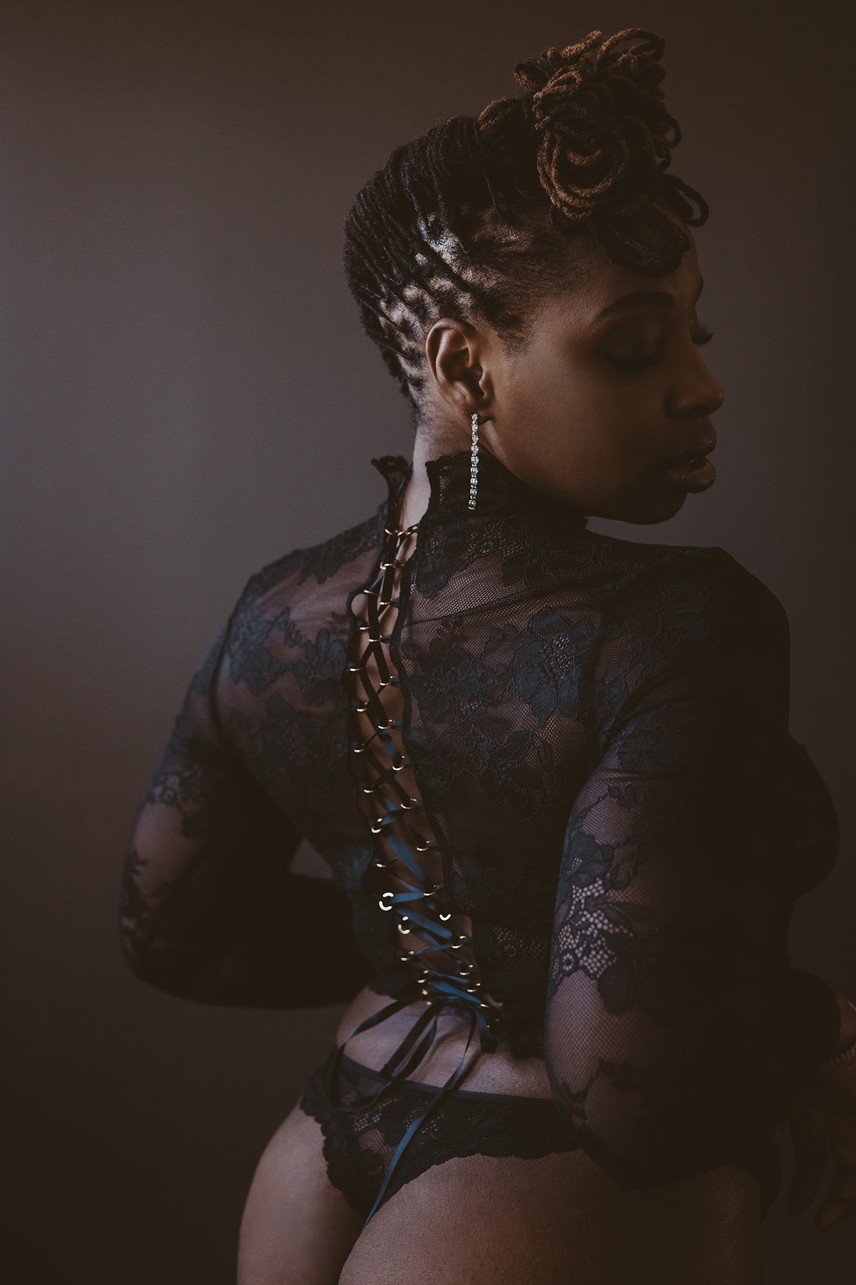 Side view of a woman with braided hair and lace clothing, looking downward, against a dark background.