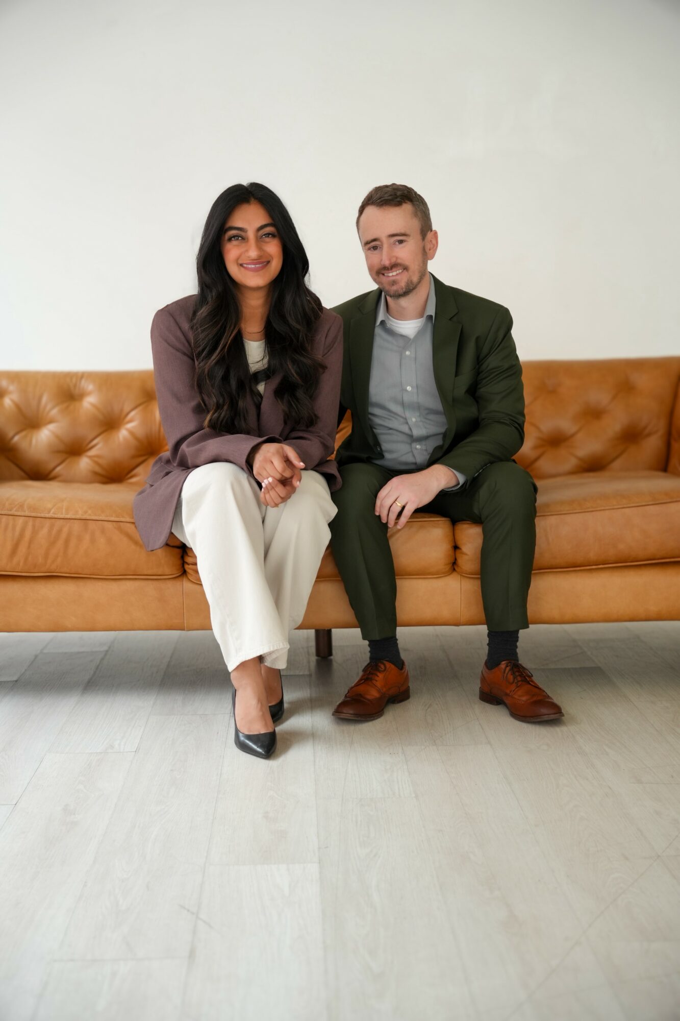 Two people sitting on a tan leather sofa, smiling, with a plain white wall background.