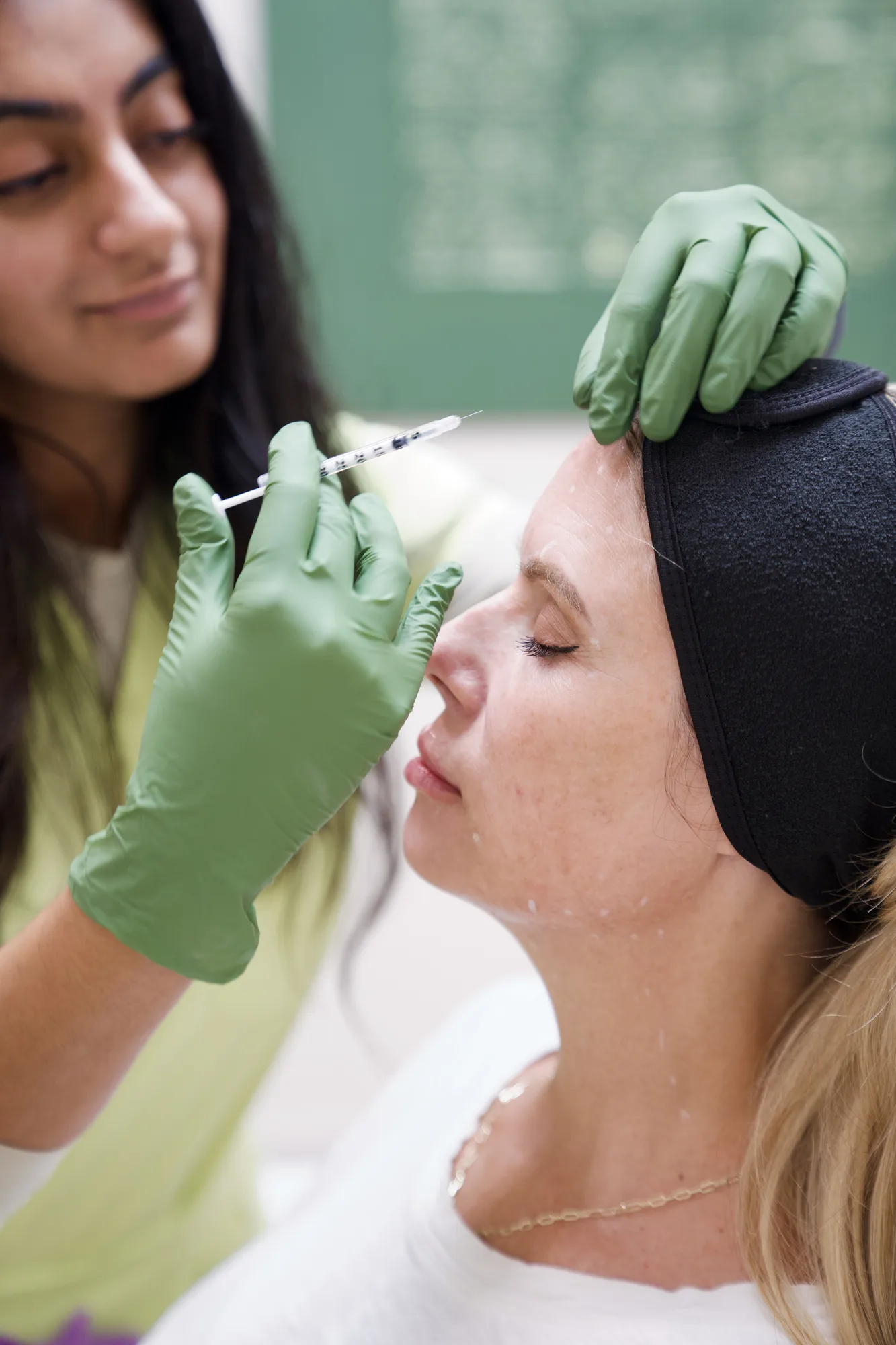 A woman receives a cosmetic injection in her forehead from a healthcare professional wearing green gloves.