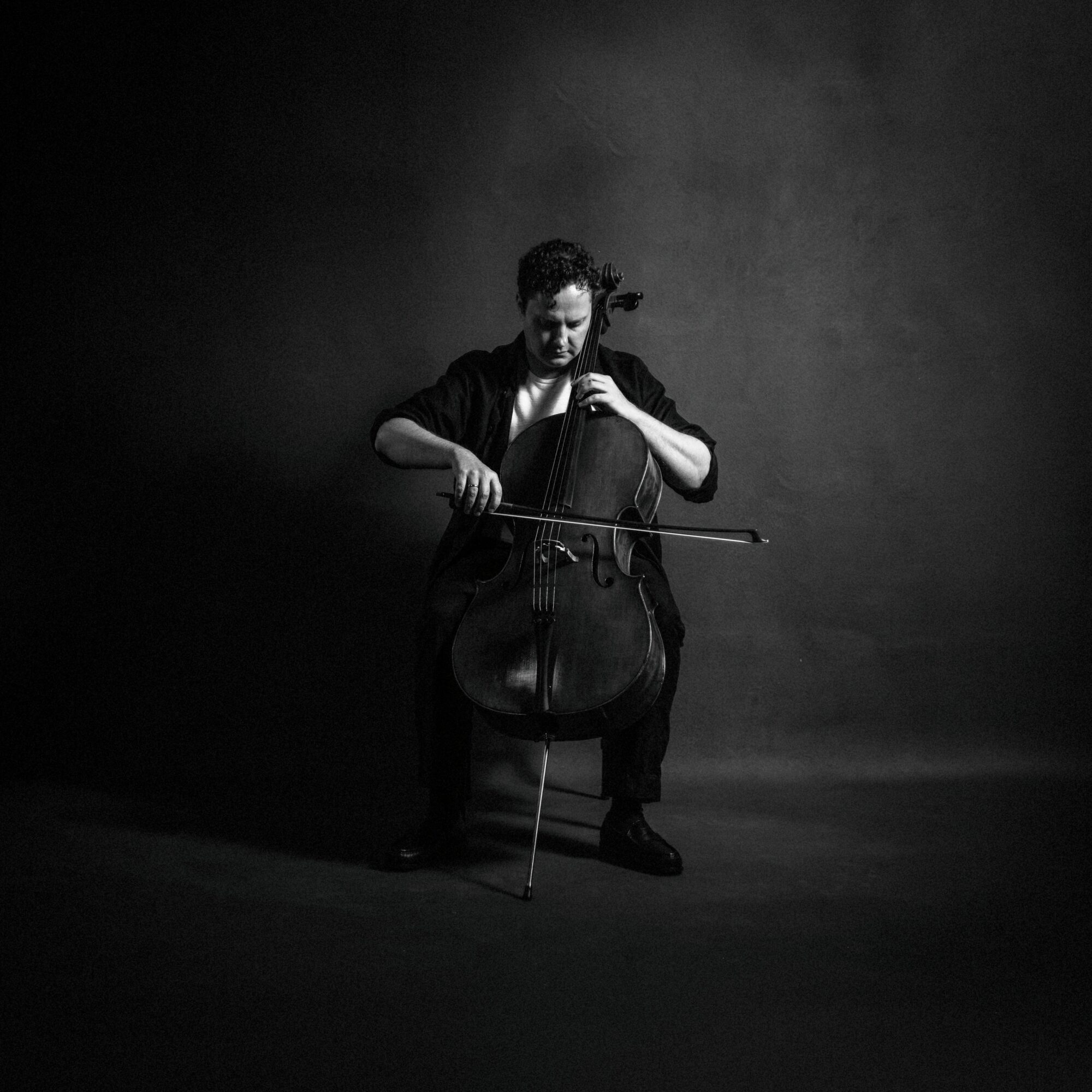 Person playing a cello in a dark setting, focused on the instrument, with a plain background.