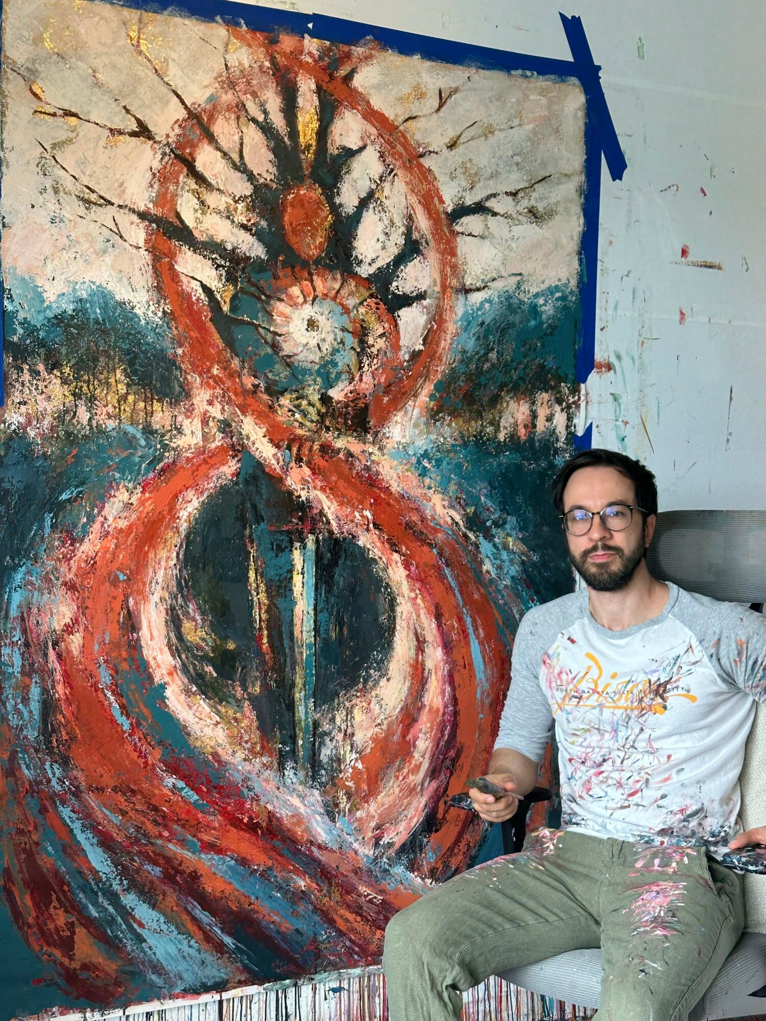 Man with glasses and beard sitting next to a large abstract painting with spiral and tree-like shapes.