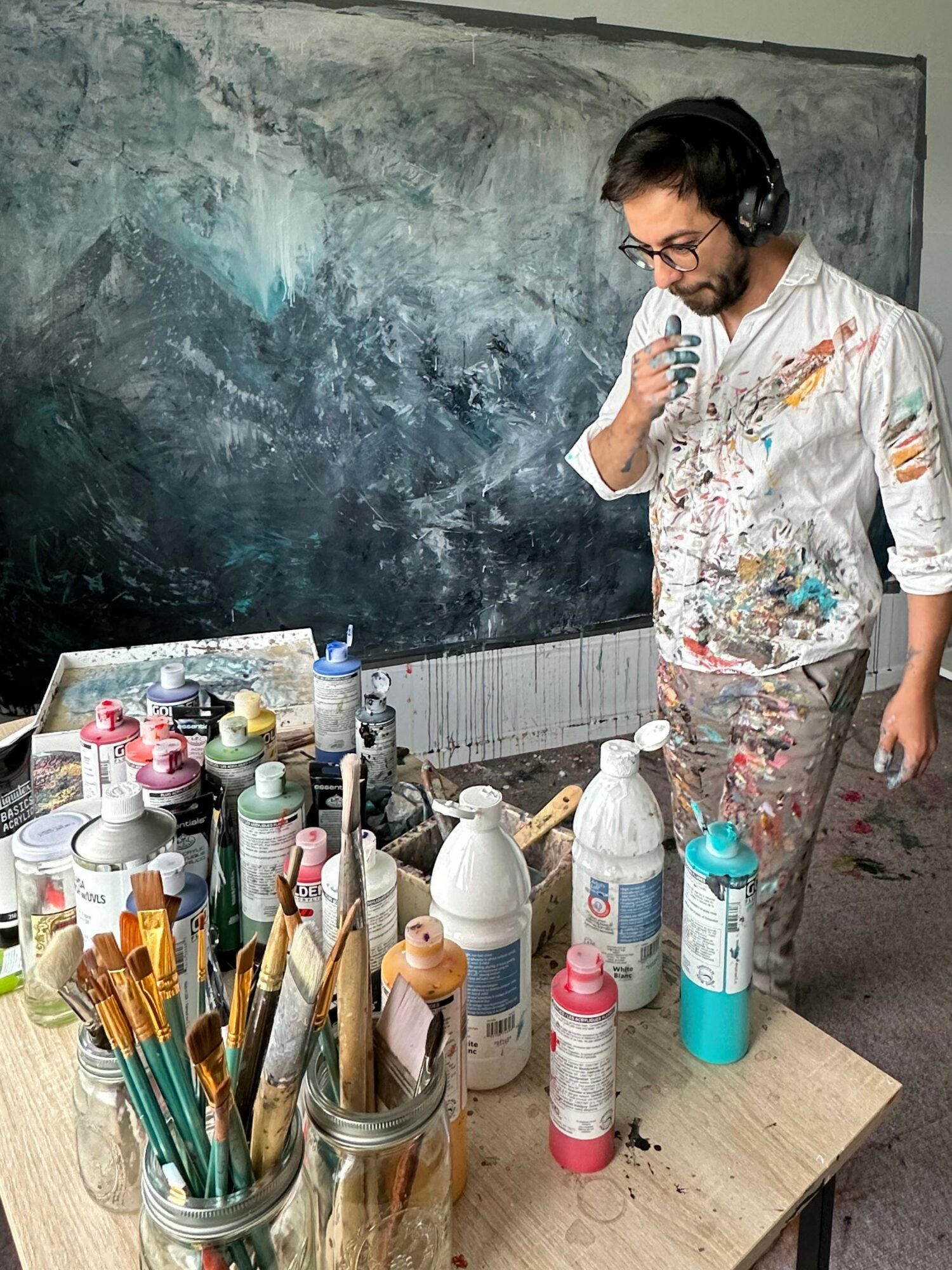 Person with glasses and paint-splattered clothes looking at a small object in hand, standing near a table with art supplies, blackboard in background.