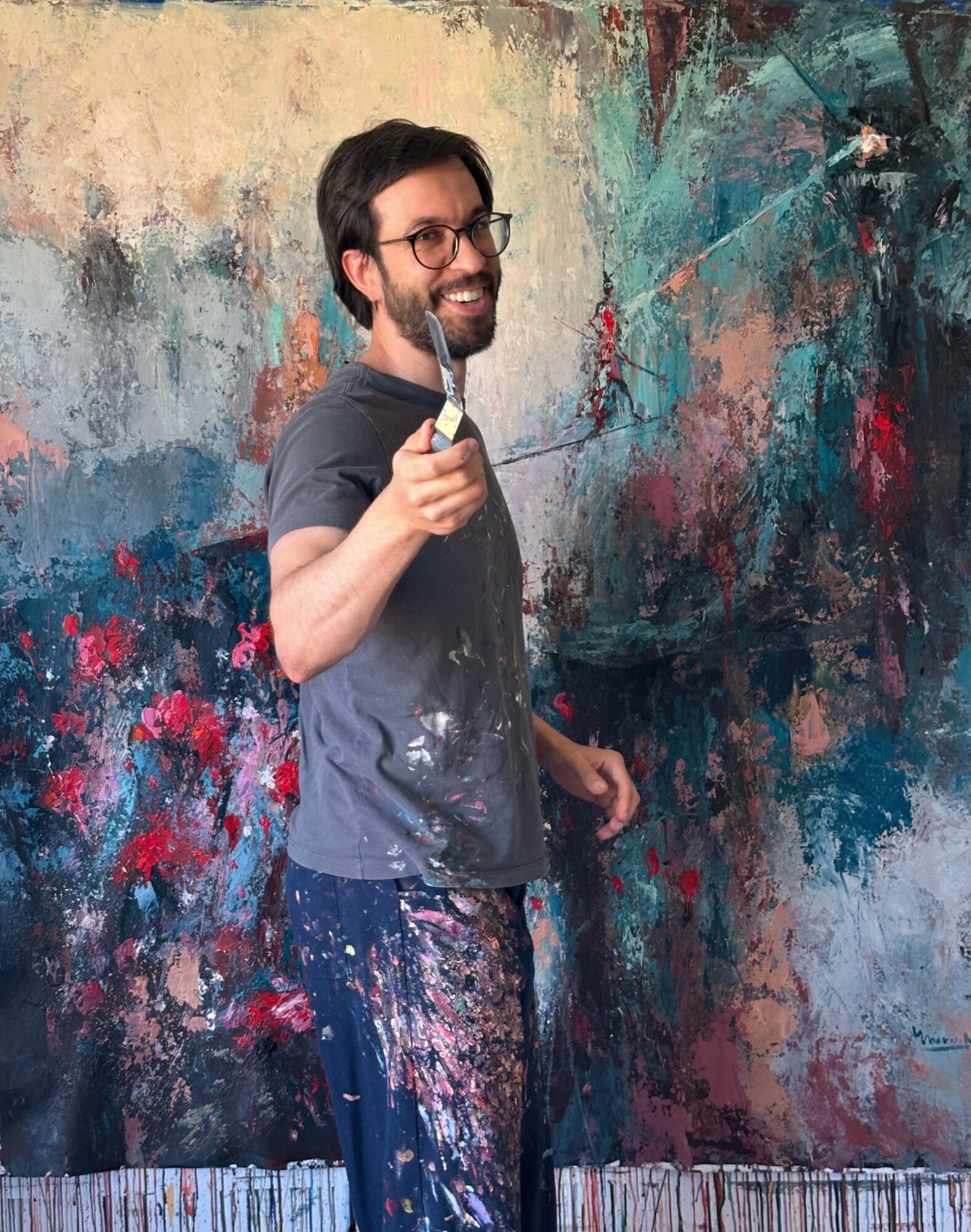 Man with glasses and beard smiling, holding a paintbrush, standing in front of an abstract colorful wall.