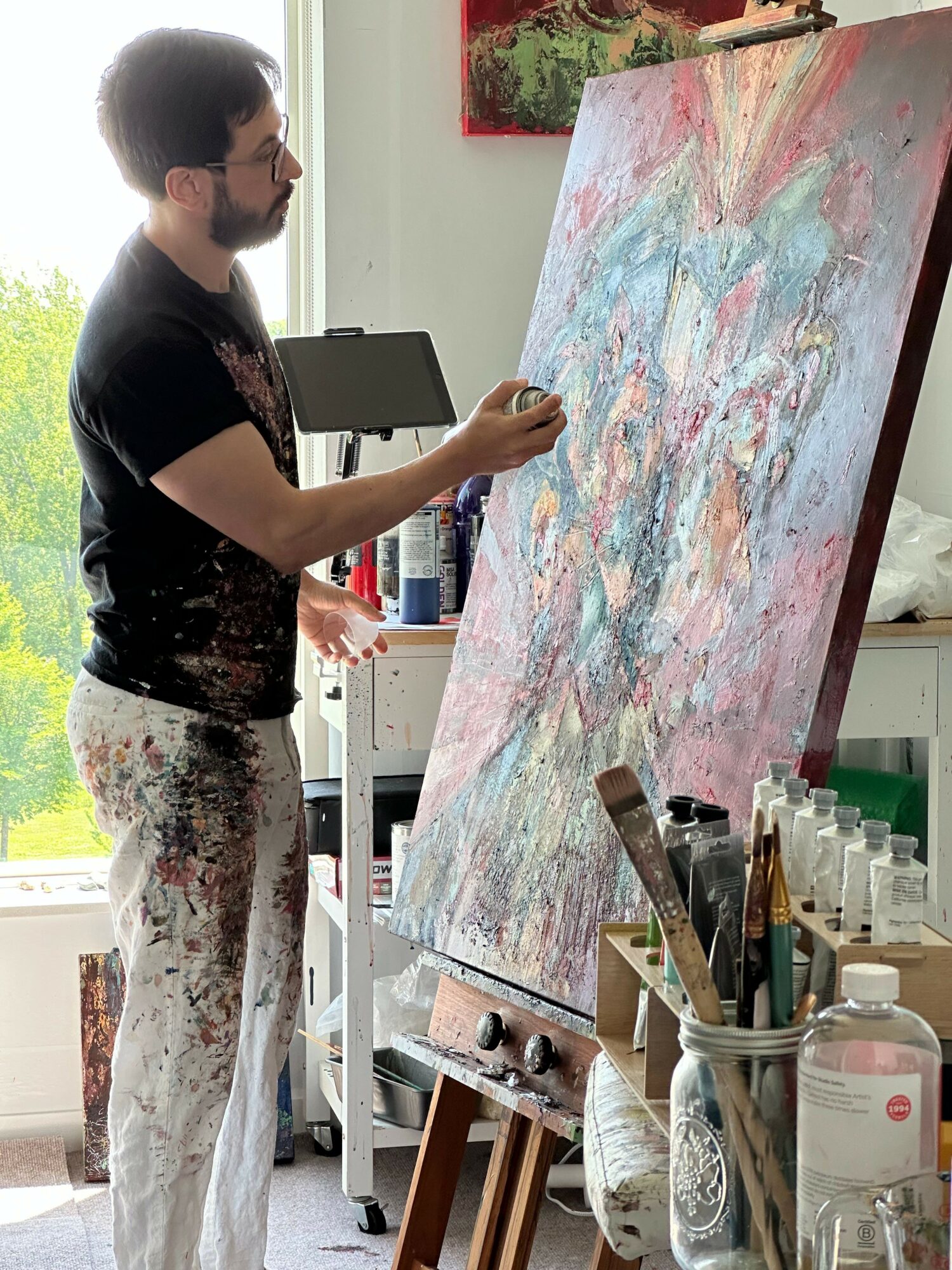 Person standing and painting on a large canvas in an art studio, with art supplies and shelves in the background.