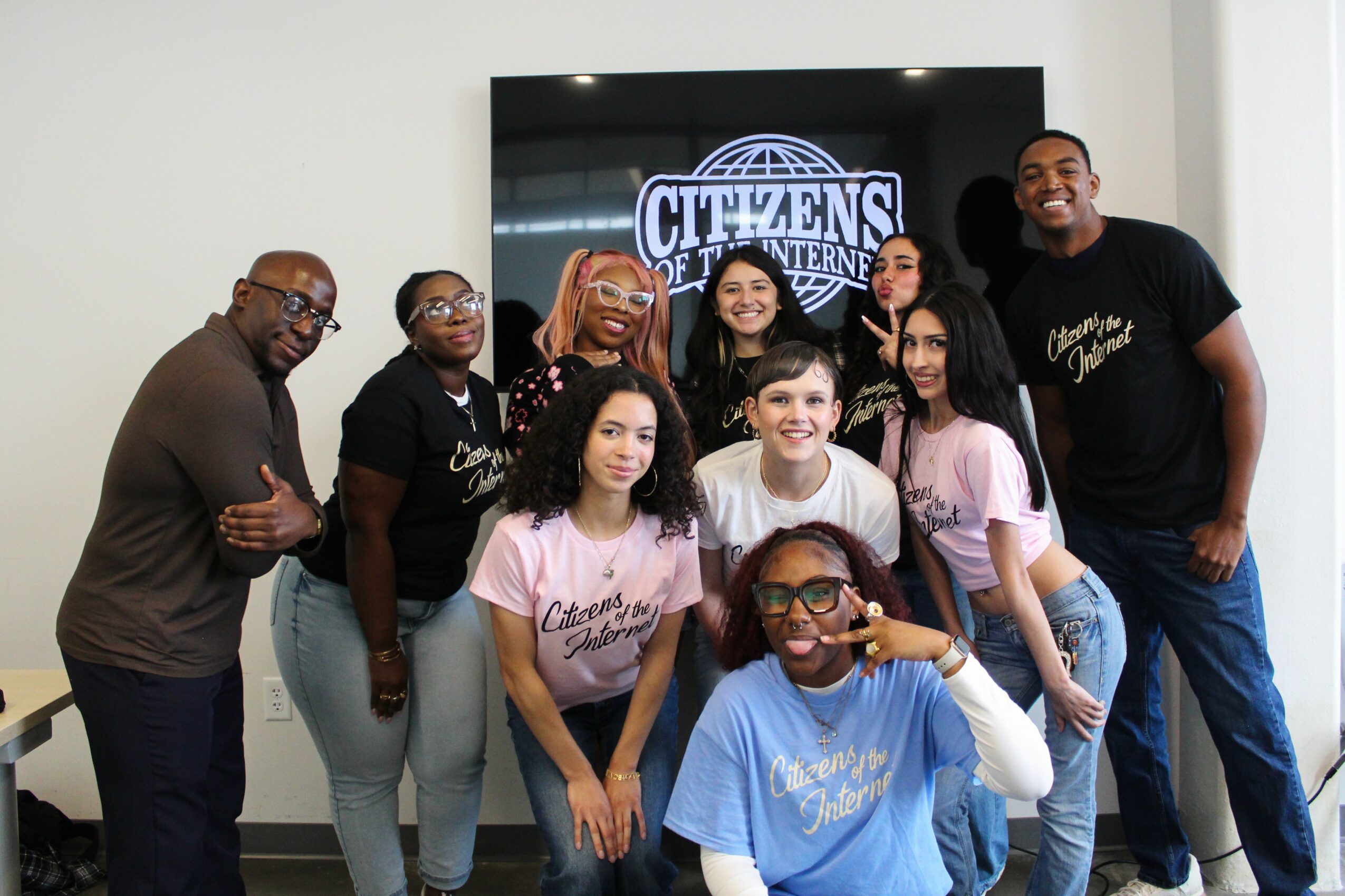 Group of nine people, some wearing pink and blue shirts, standing in front of a screen with a logo, smiling.