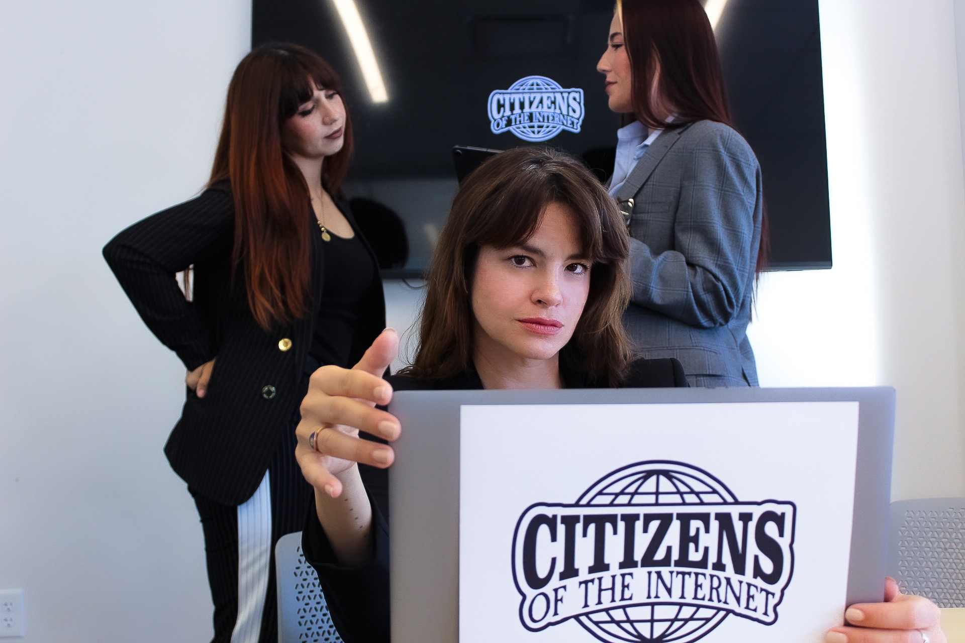 Three women in a modern office, one holding a sign that reads 'CITIZENS OF THE INTERNET,' with a screen in the background.