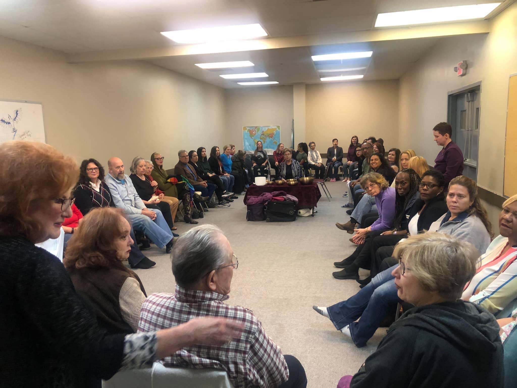 People seated in a large circle in a room, participating in a group discussion or meeting.