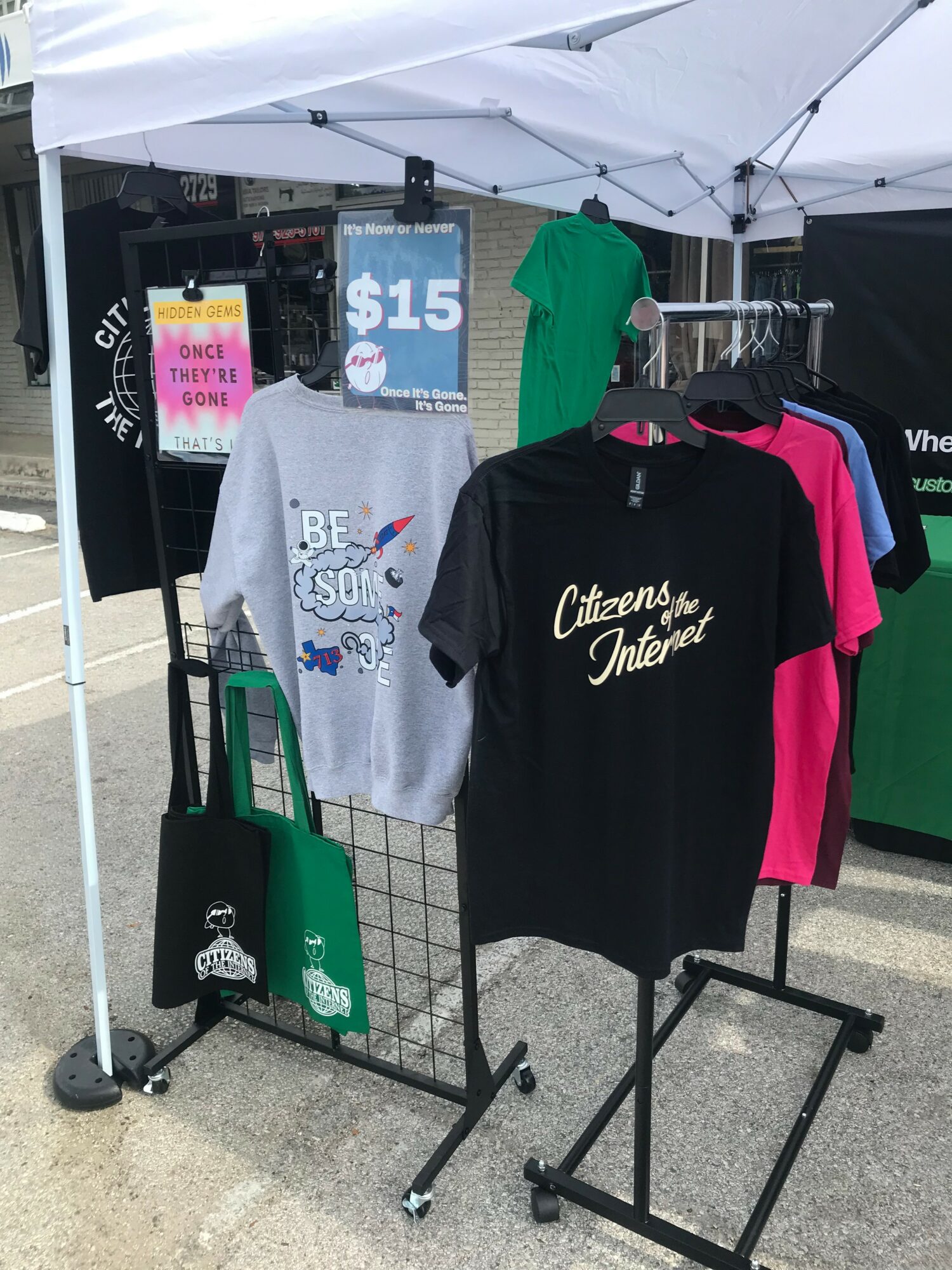 Clothing display with gray and black shirts, a green shirt in the background, and a sign showing $15 price under a white canopy.