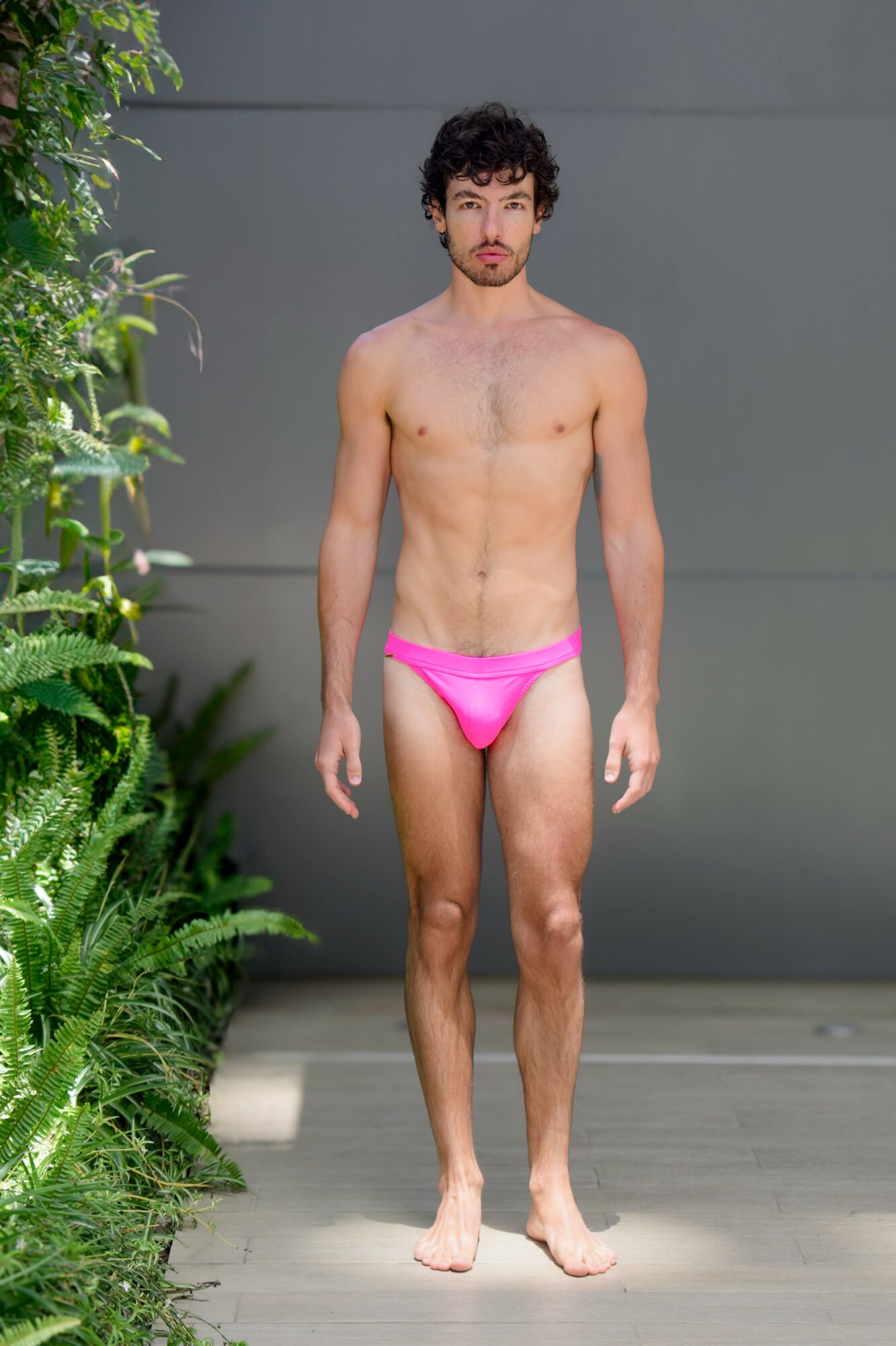 Young man with curly hair and a beard stands outdoors in pink swimwear, facing forward, with greenery on the left.