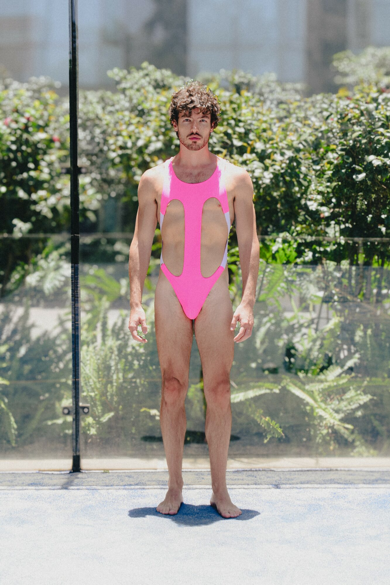 Young man in pink swimsuit standing outdoors with greenery background, facing forward, arms at sides, sunlight, and shadows visible.