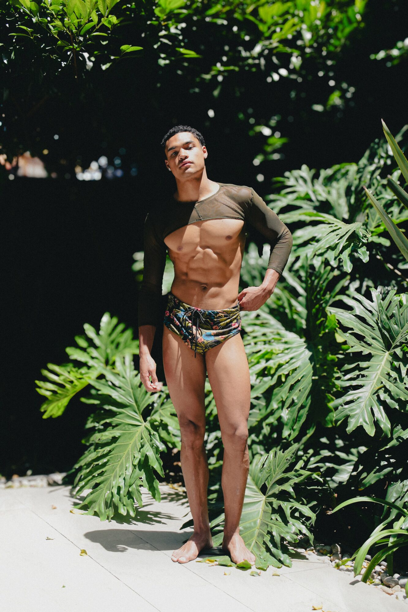 Woman standing outdoors among large green tropical plants, wearing a bikini and a sheer cover-up, with sunlight on her face and body.