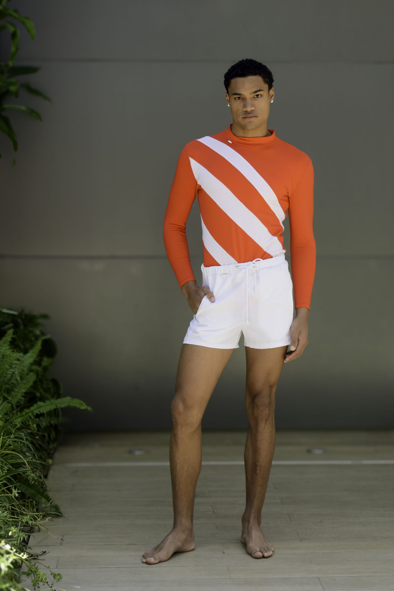 Young man standing barefoot in front of a plain wall, wearing a red and white striped long-sleeve shirt and white shorts.