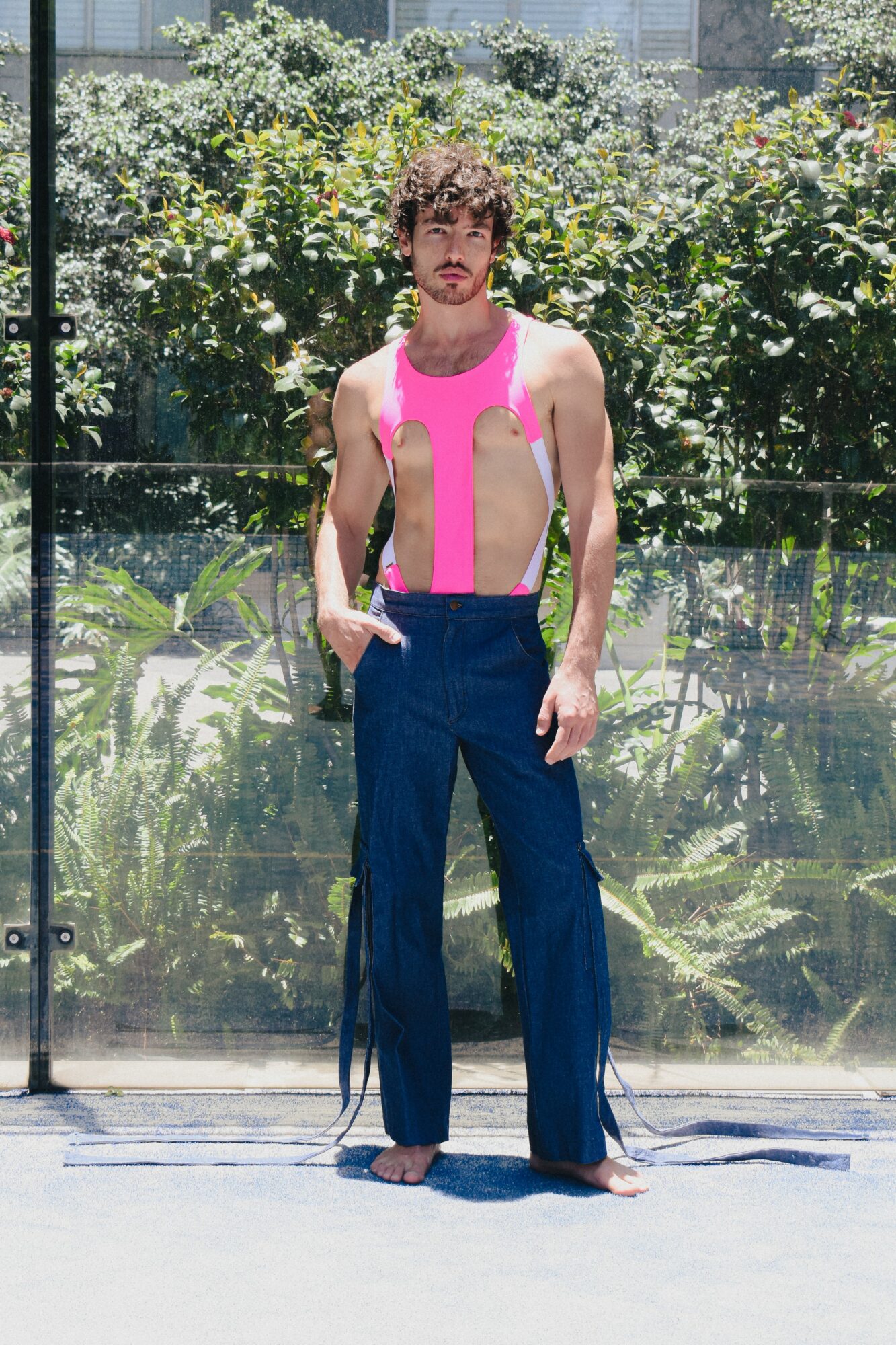 Person standing outdoors with greenery behind, wearing a pink and white top and dark blue pants, barefoot.