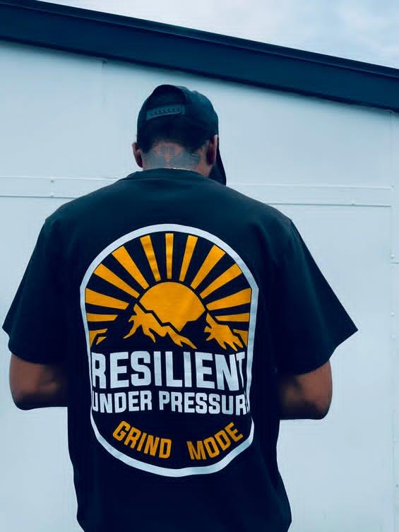 Person wearing a black T-shirt with a graphic design of a sun, mountains, and the words 'Resilient Under Pressure' and 'Grind Mode'.