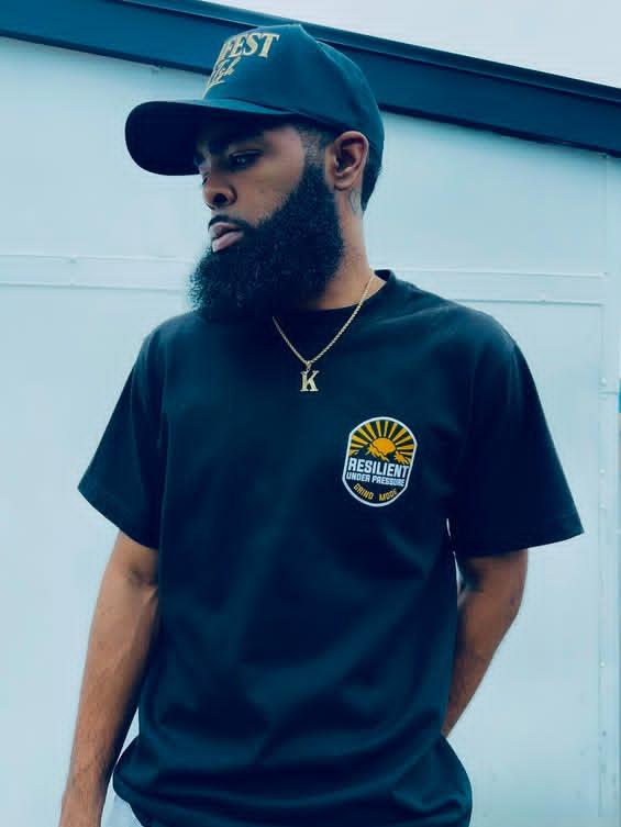 Young man with a beard wearing a baseball cap, black T-shirt with a logo, and a gold necklace with the letter K, standing outdoors.