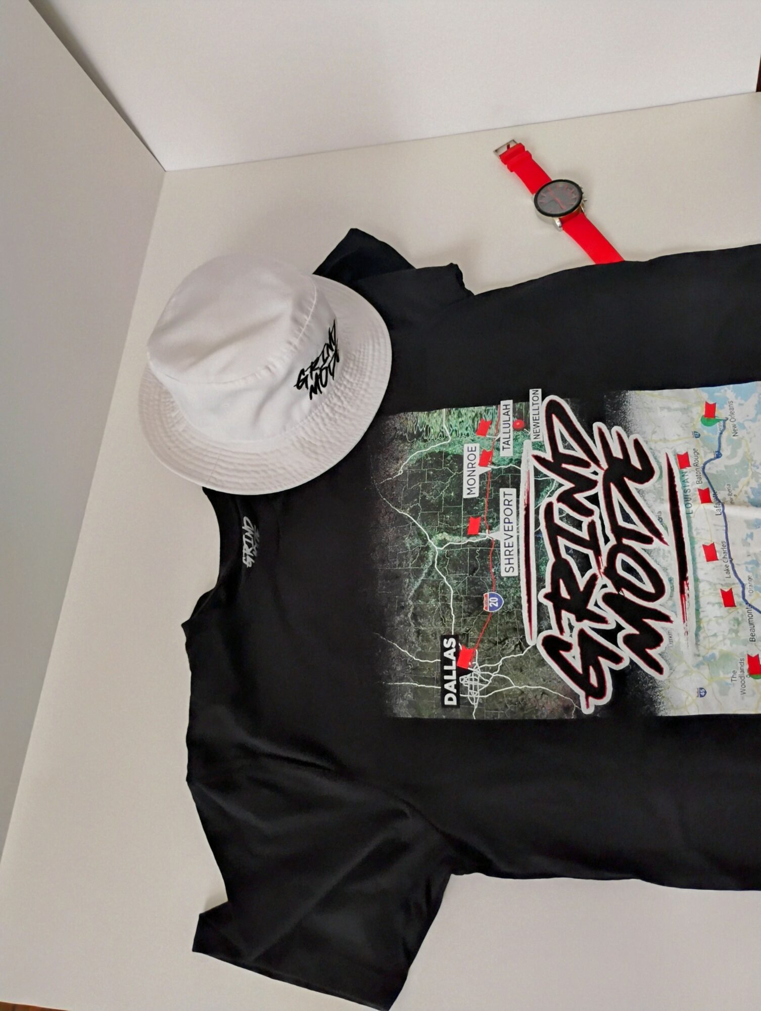 Black T-shirt with graphic design, white hat, red watch, and black cloth on a white surface.