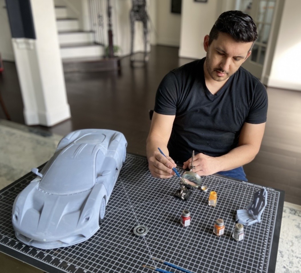 Man painting a model car on a black grid surface in a room with wooden floors and white walls.