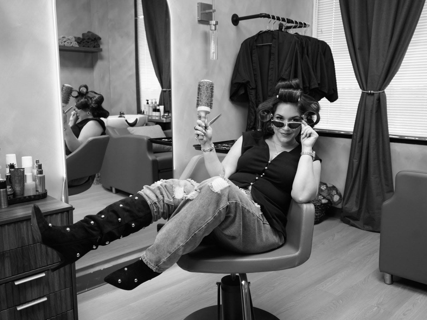Woman with hair rollers, sunglasses, and casual clothing relaxing on a chair in a room with curtains and shelves.