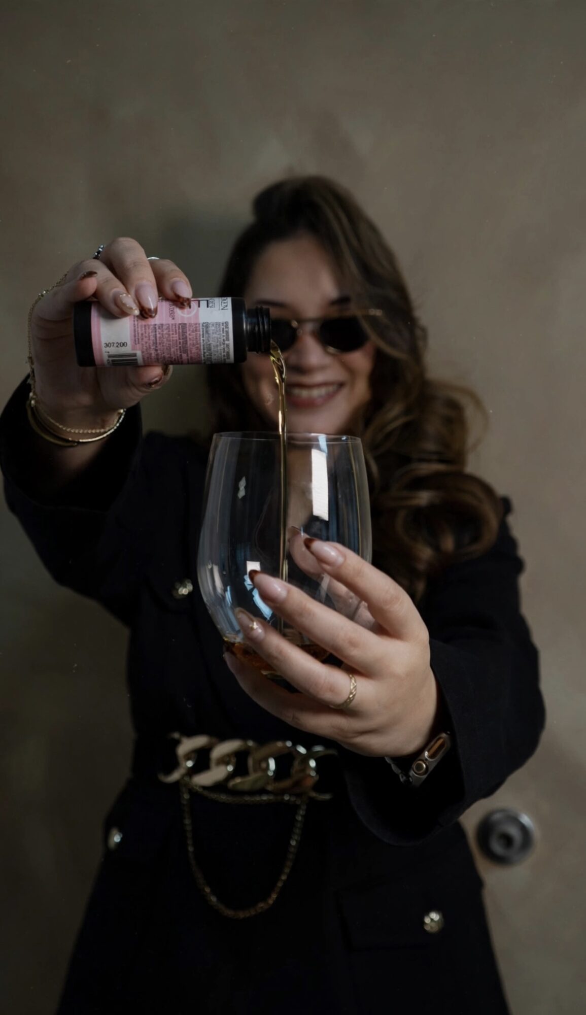 Person pouring a drink into a glass, smiling, wearing sunglasses and a dark outfit.