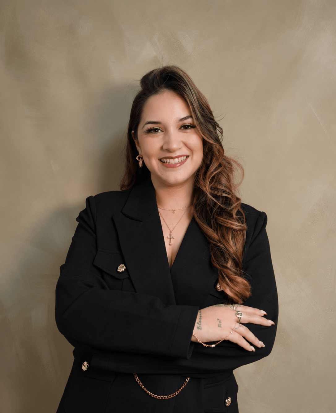 Young woman with long wavy hair smiling, wearing a black blazer, standing against a beige background.