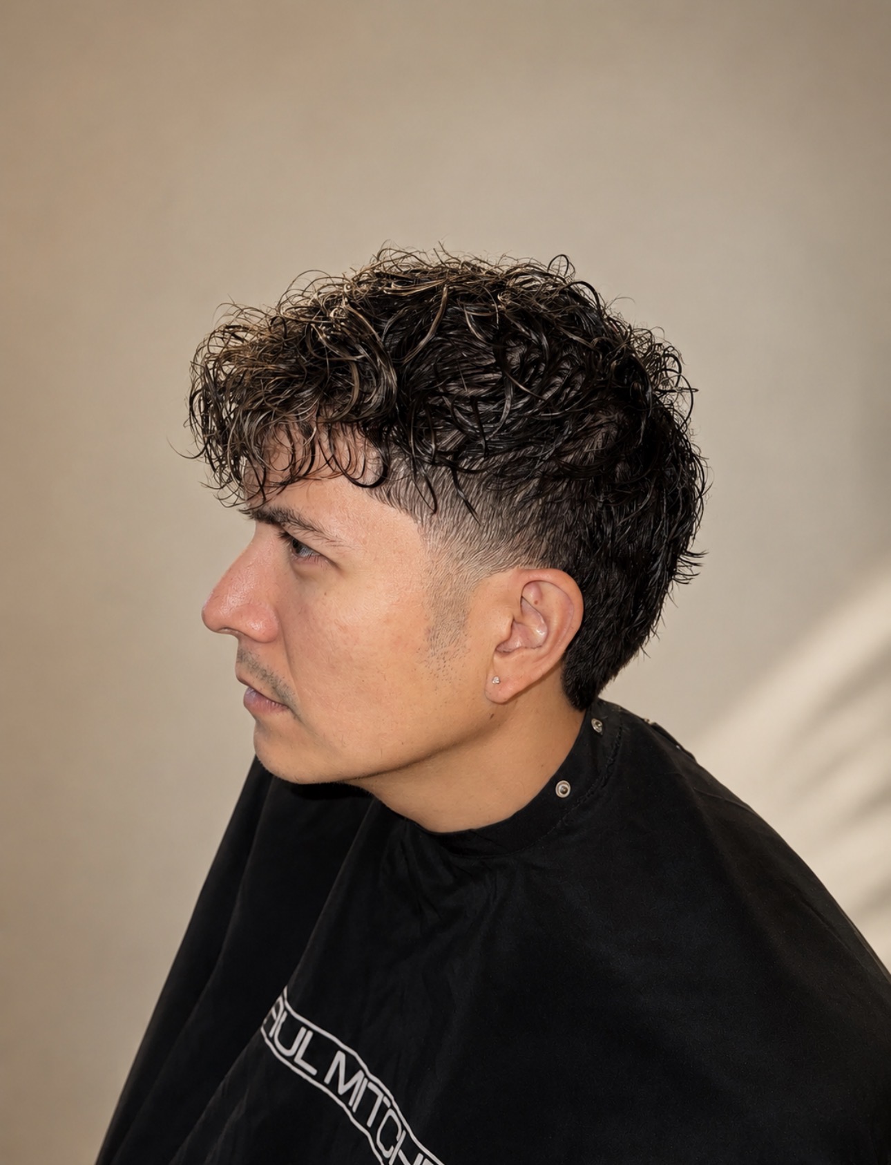 Side profile of a young man with curly hair, wearing a black shirt, against a plain background.