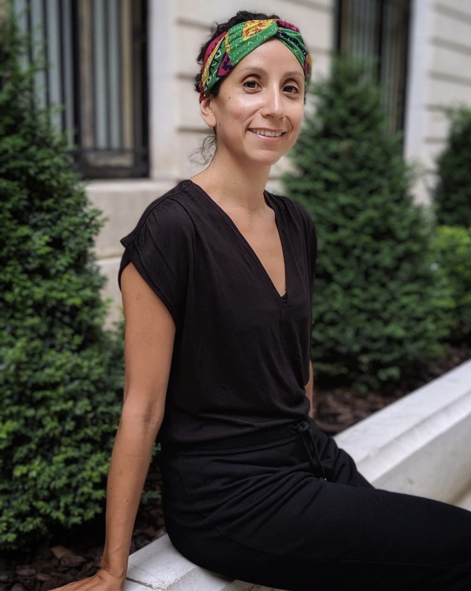Smiling woman with a colorful headscarf, black top, and black pants outdoors with greenery behind her.