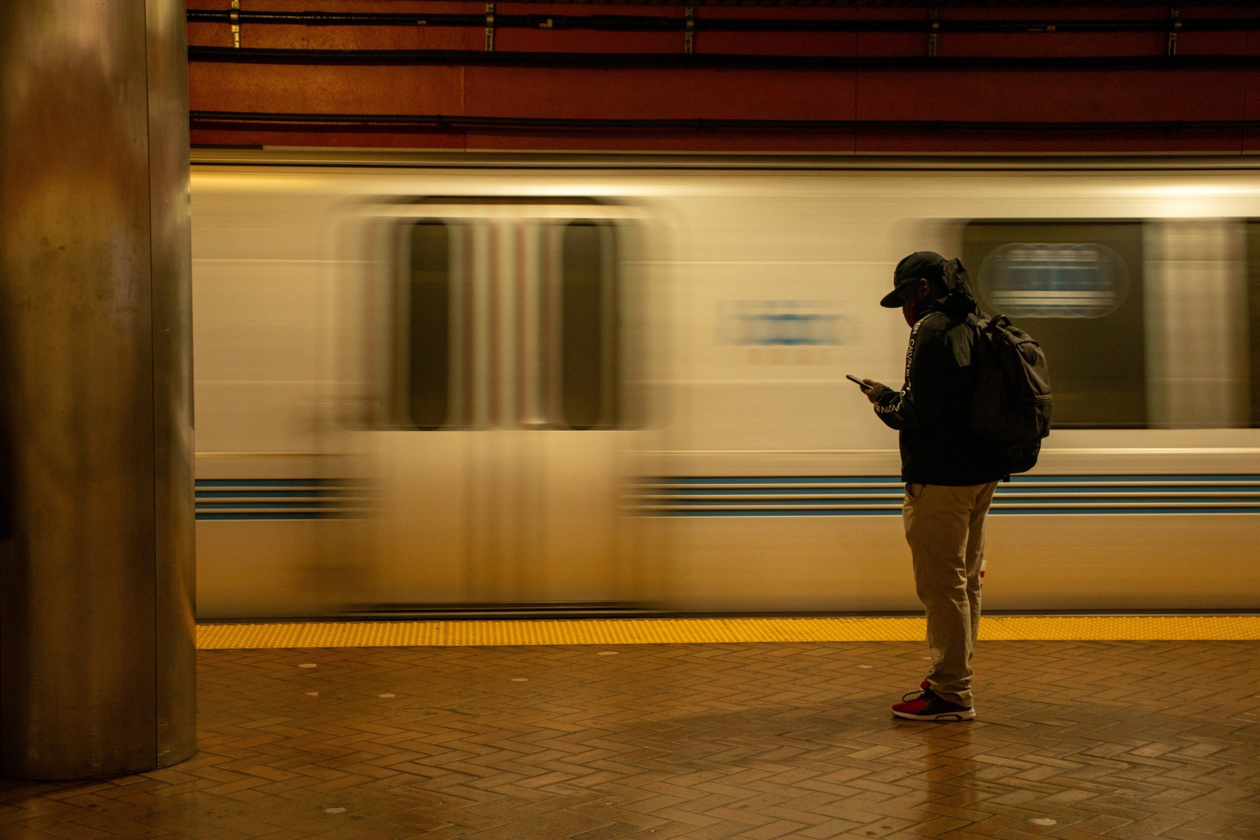 Person standing on subway platform looking at phone with moving train in background.