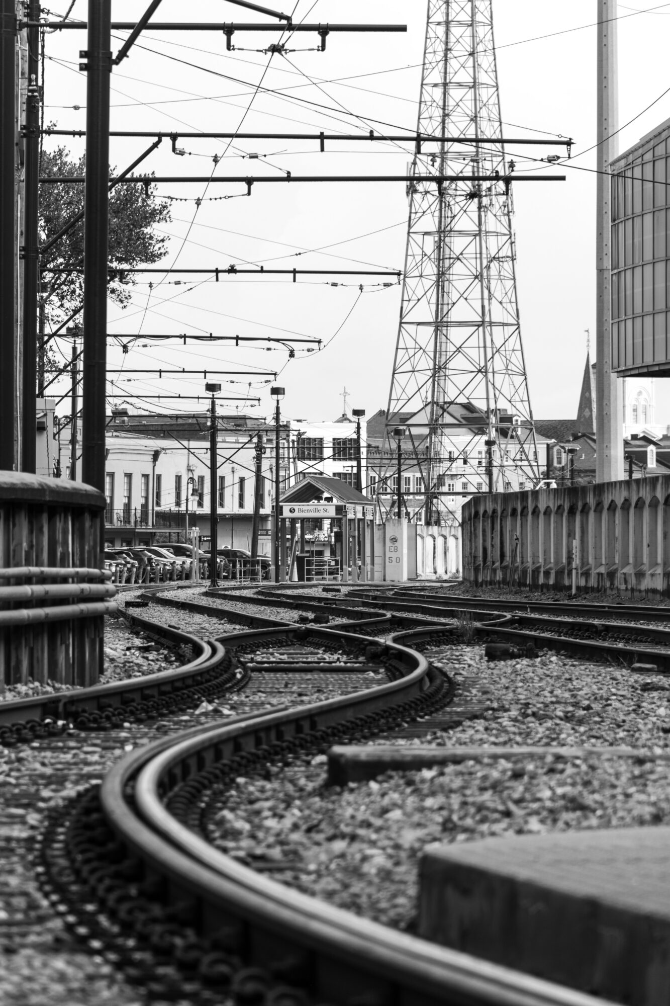 Railway tracks with overhead wires and a large electrical tower in an urban setting, black and white.