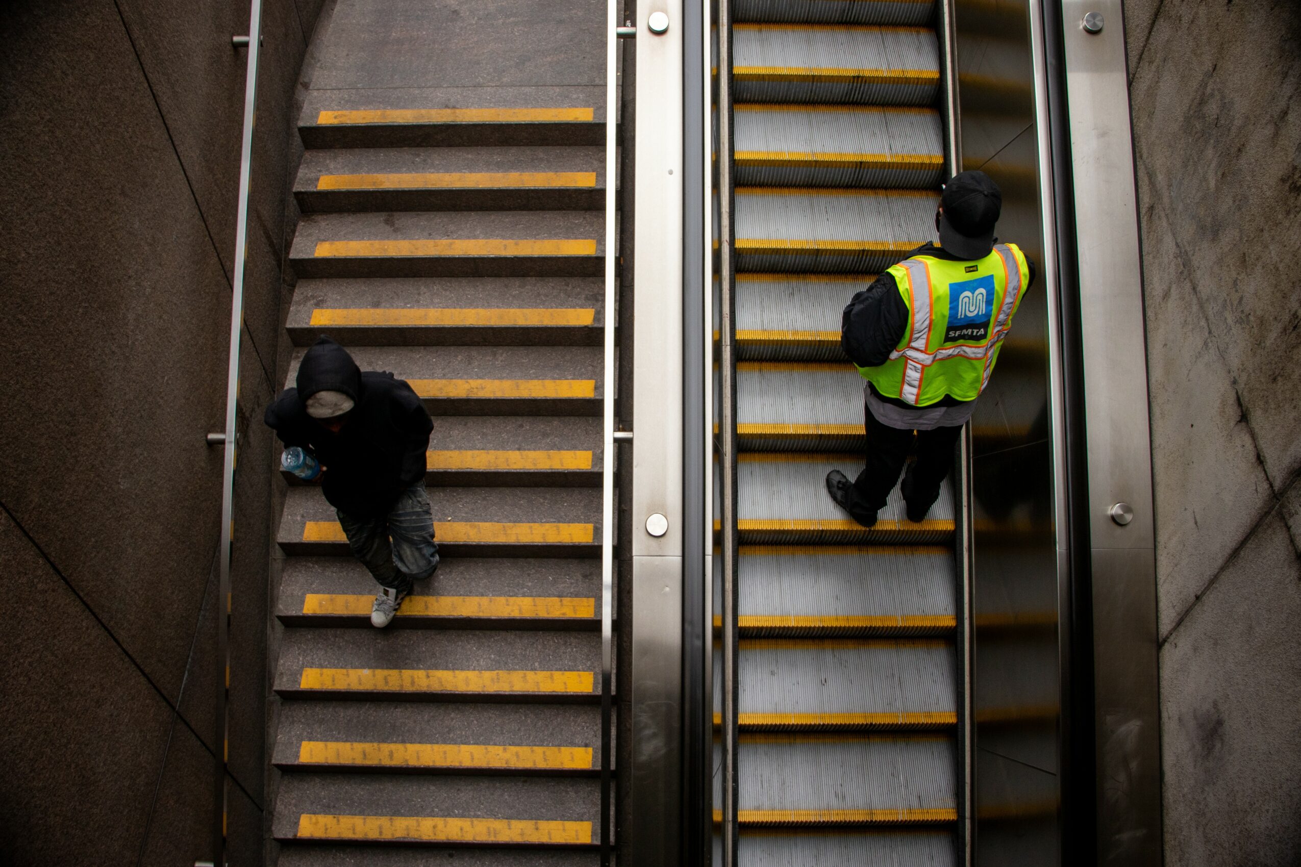 Two people on separate escalators, one ascending and one descending, in an indoor setting with concrete walls.