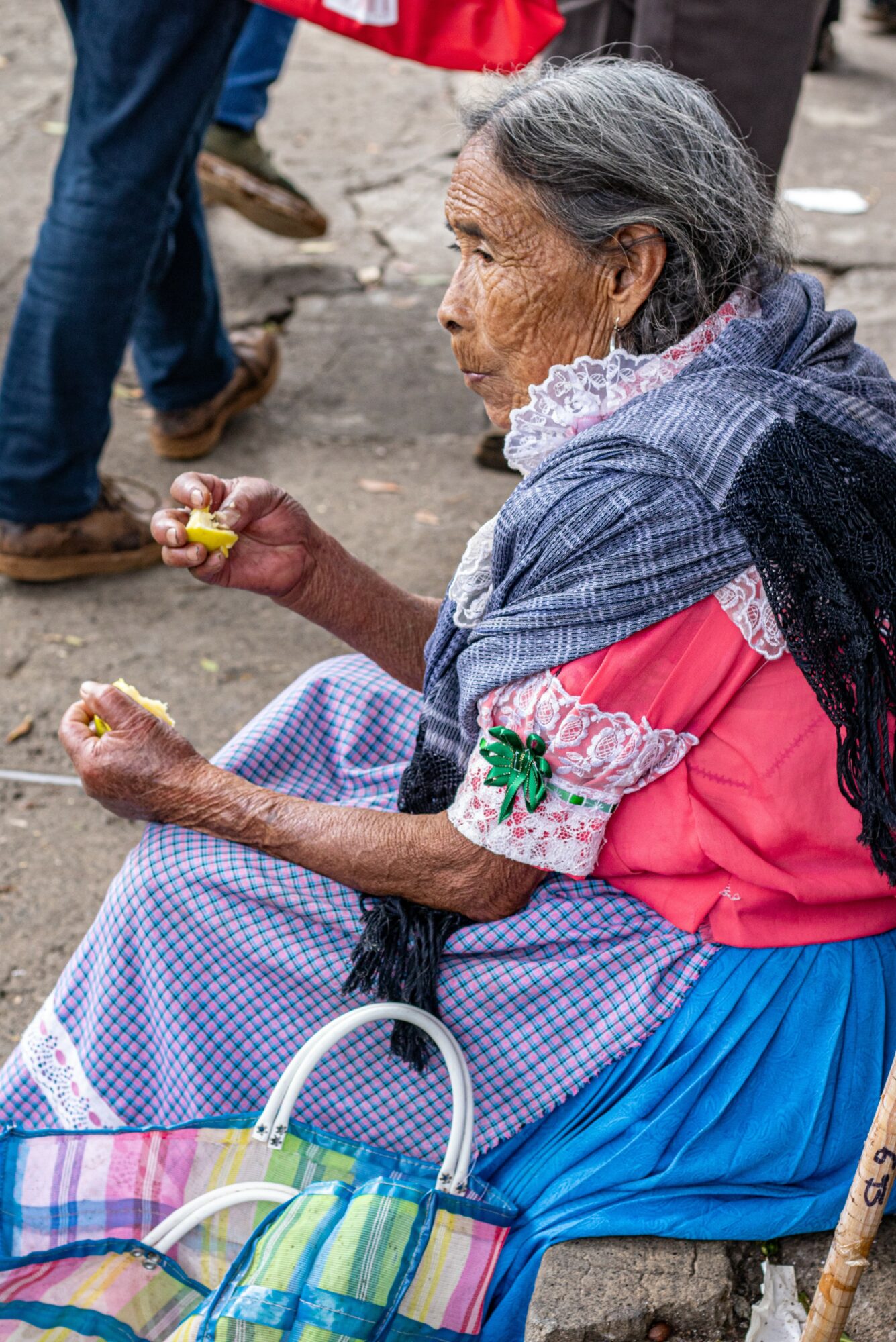 Elderly woman sitting outdoors, holding a small object, wearing a pink top with lace and a blue skirt, with a colorful bag nearby.