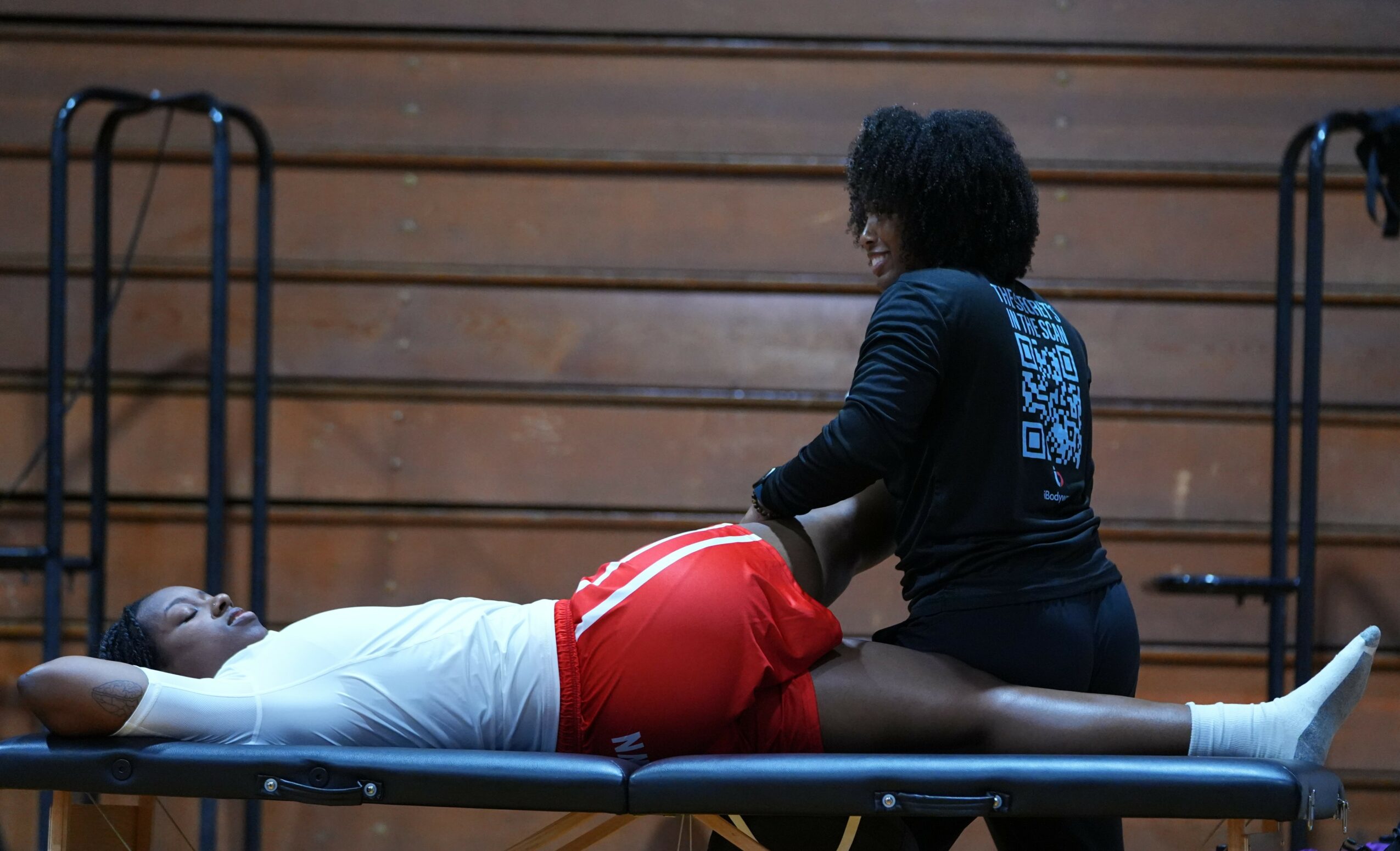 Person lying on a table receiving treatment from a healthcare worker in a gymnasium.