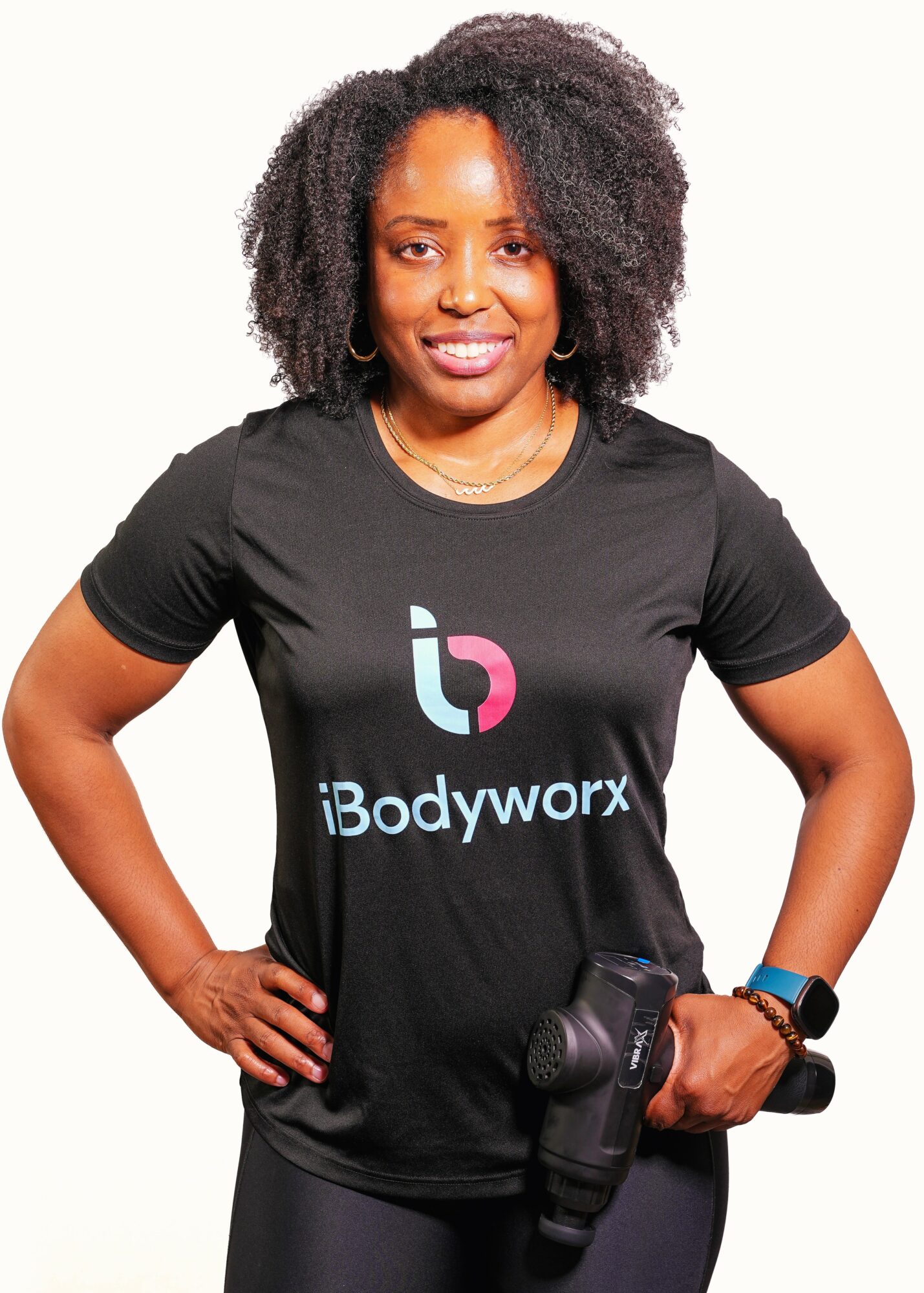 Woman with curly hair wearing a black t-shirt with a logo, holding a camera, smiling, standing with hands on hips.