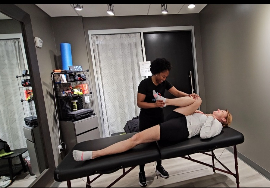 Person lying on a massage table receiving treatment from a therapist in a room with gray walls and a sliding door.