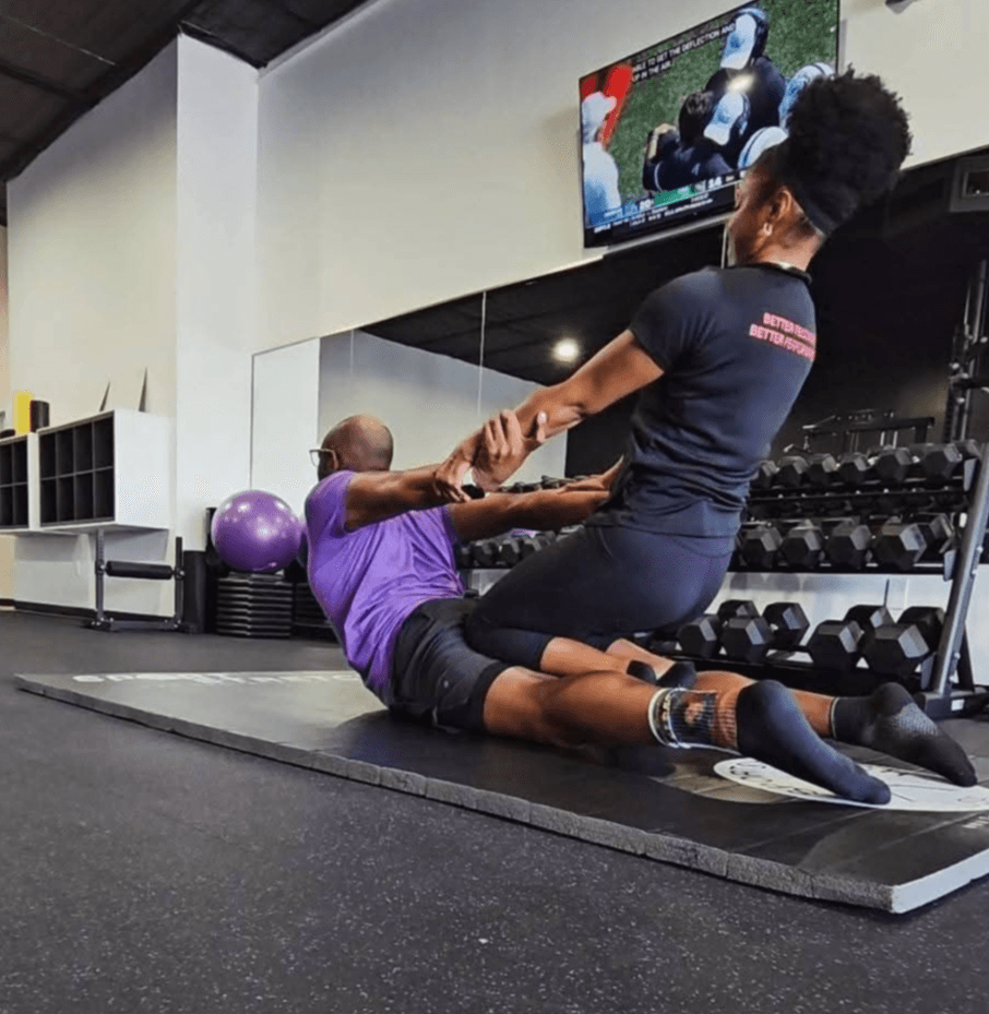 Two people exercising on a mat in a gym, one assisting the other with stretching, with gym equipment and a TV screen in background.