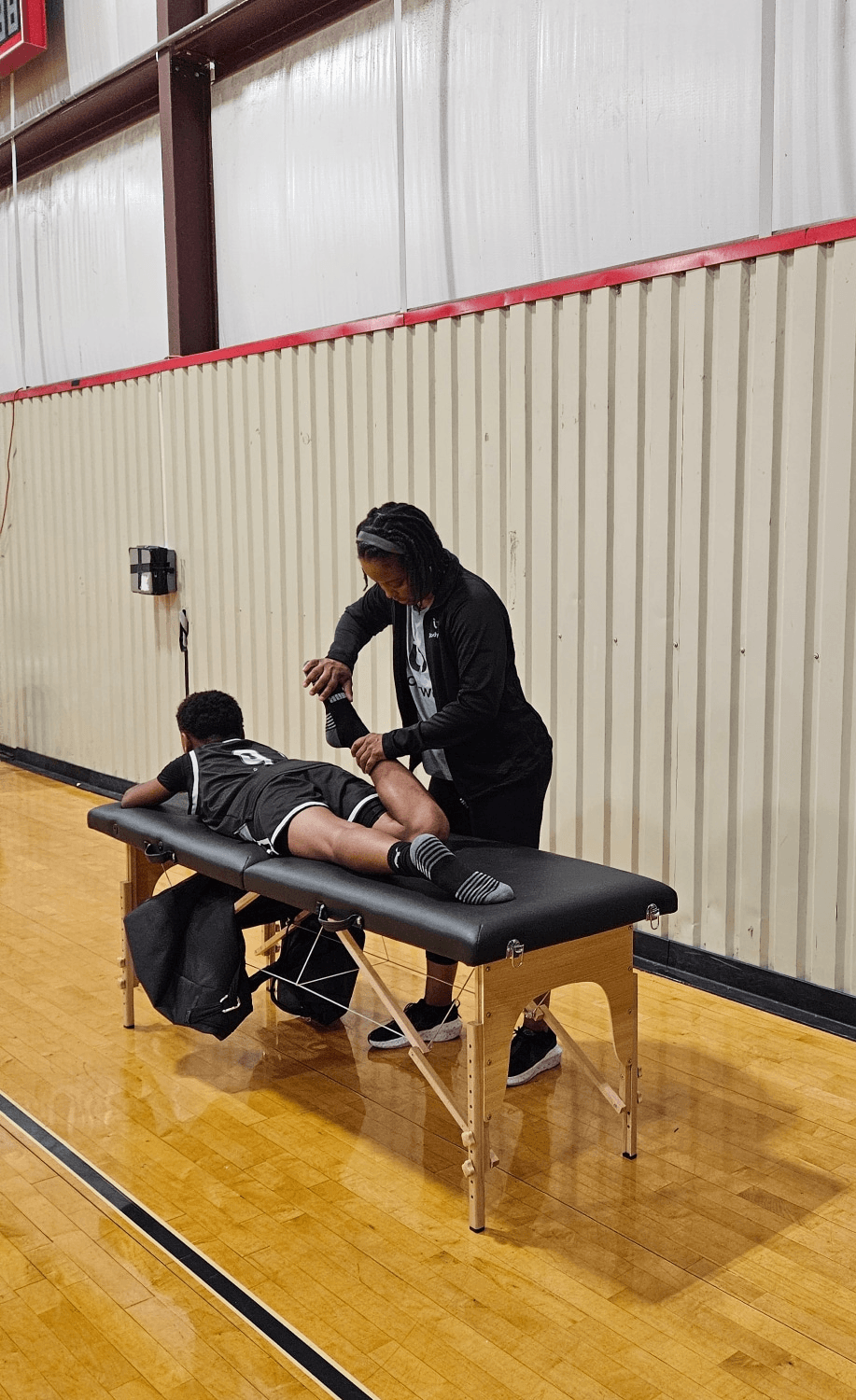 Person lying face down on a massage table with a backpack nearby, in a gymnasium with a wooden floor and metal wall.