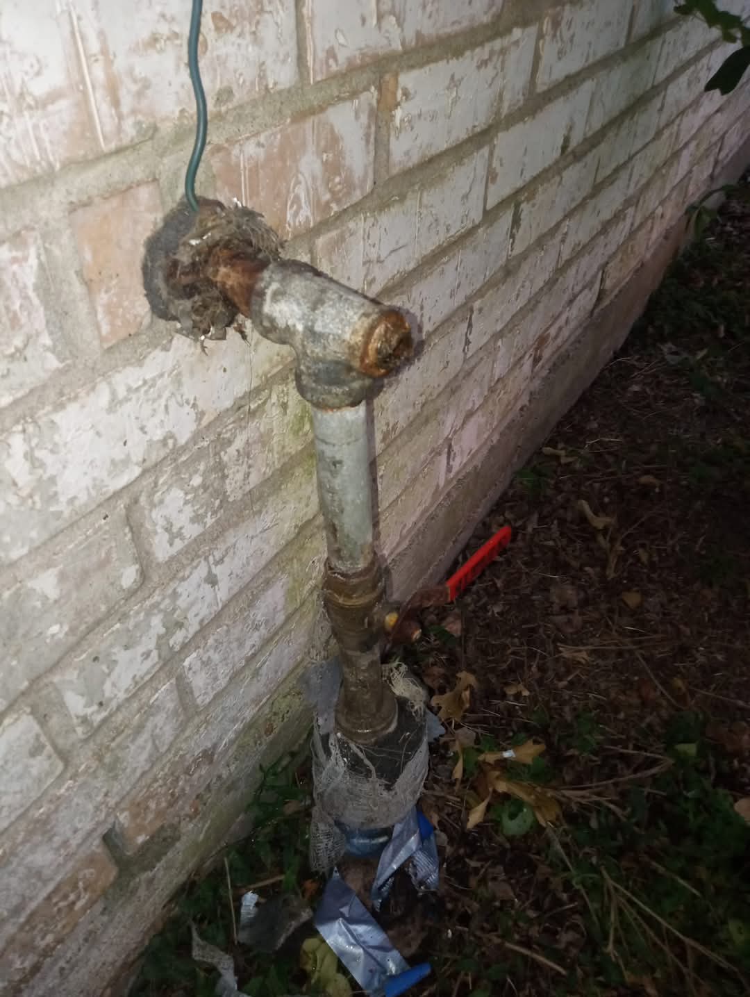 A rusty outdoor pipe with a valve attached, connected to a brick wall, with some plants and soil nearby.