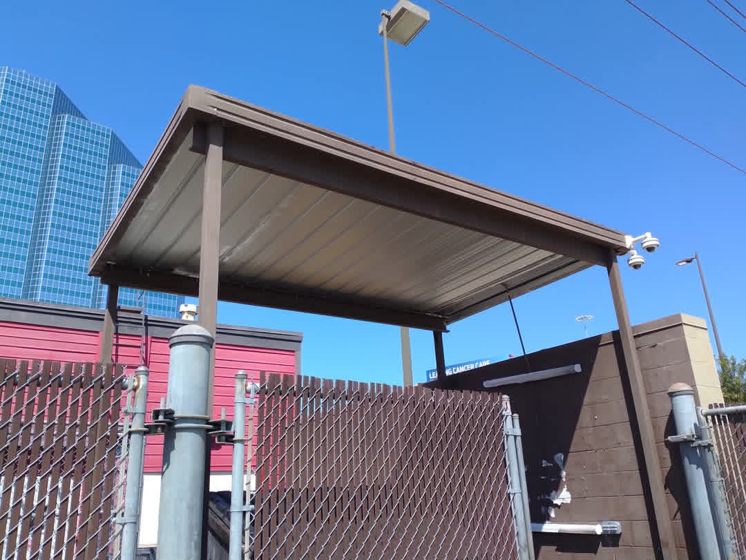 Upright view of a small outdoor shelter with a metal roof, chain-link fence, and utility equipment, against a blue sky.