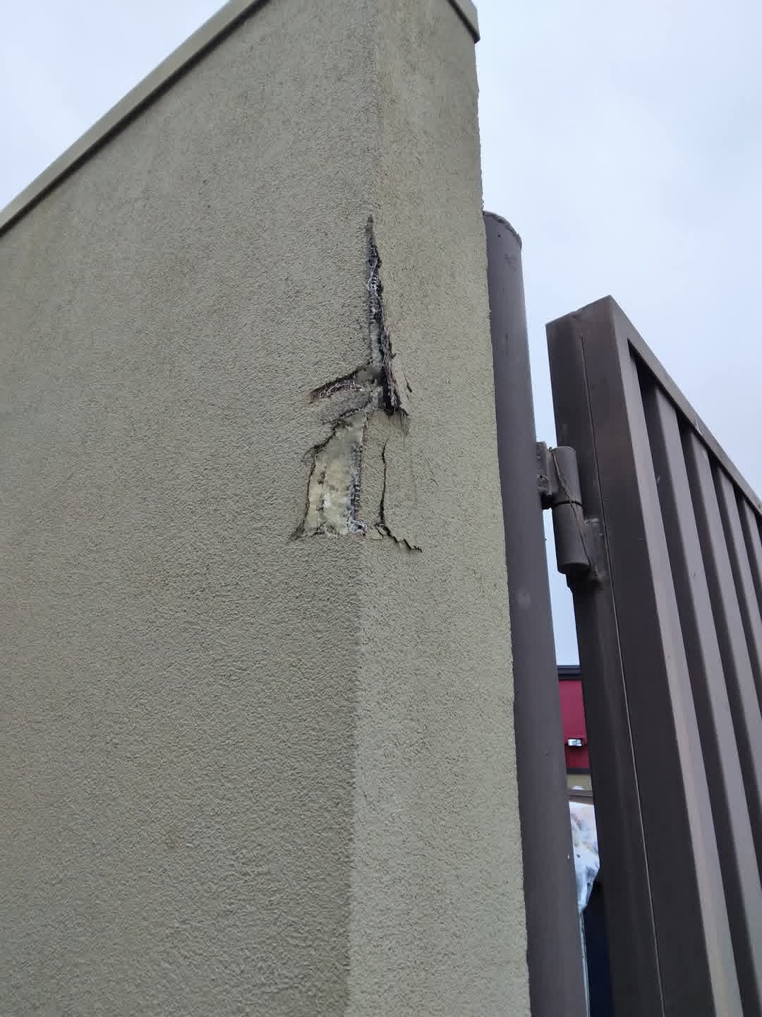 Concrete wall with a vertical crack and damage near a metal gate, against a cloudy sky.