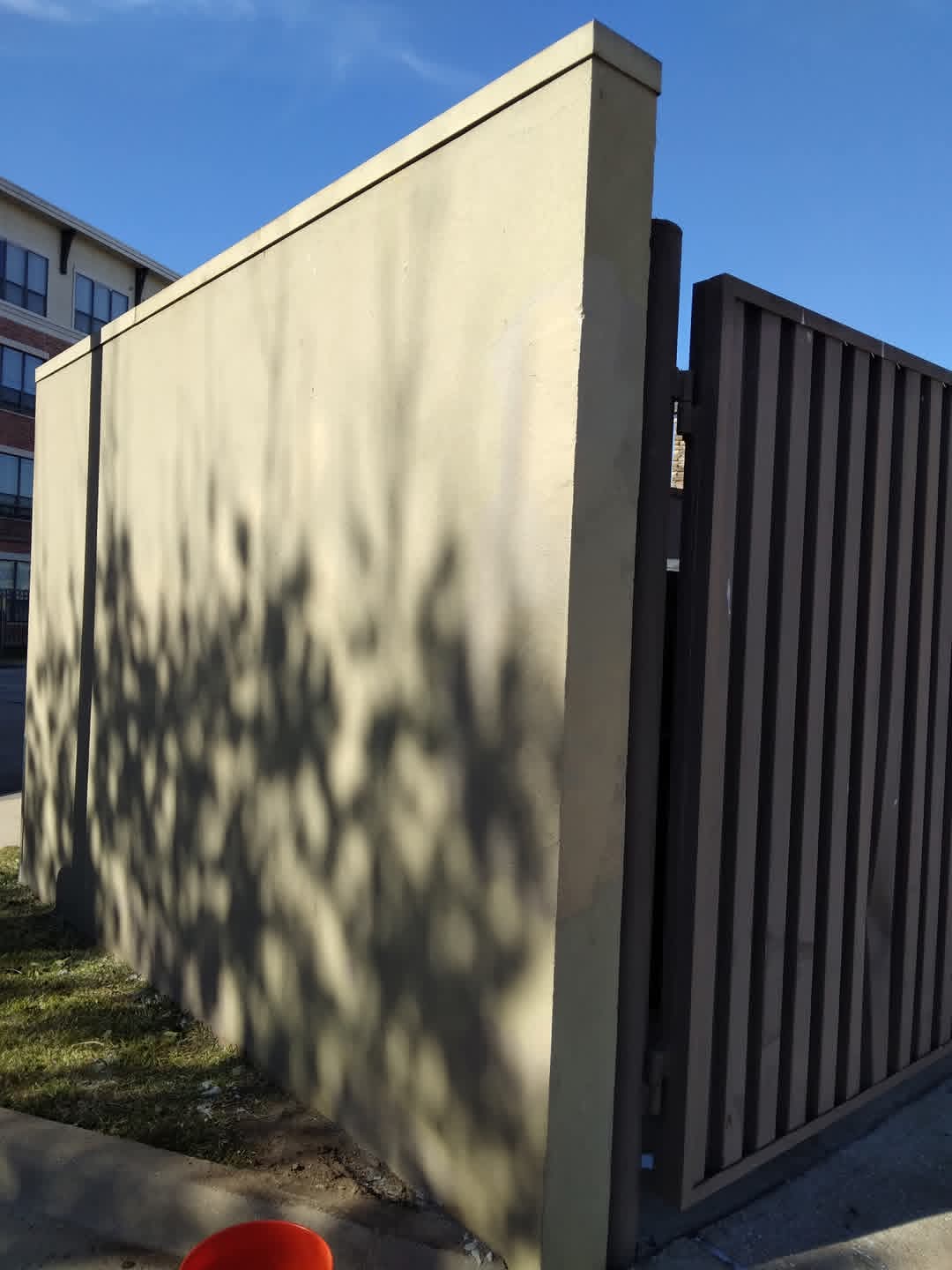 Concrete wall with shadows of trees, next to a dark metal fence, under a clear blue sky.