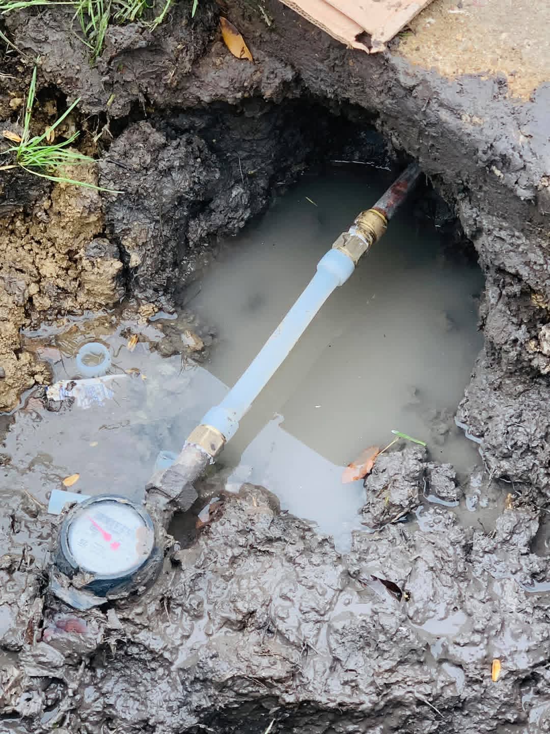 Pipe connected to a water meter in a muddy hole with water and soil around.