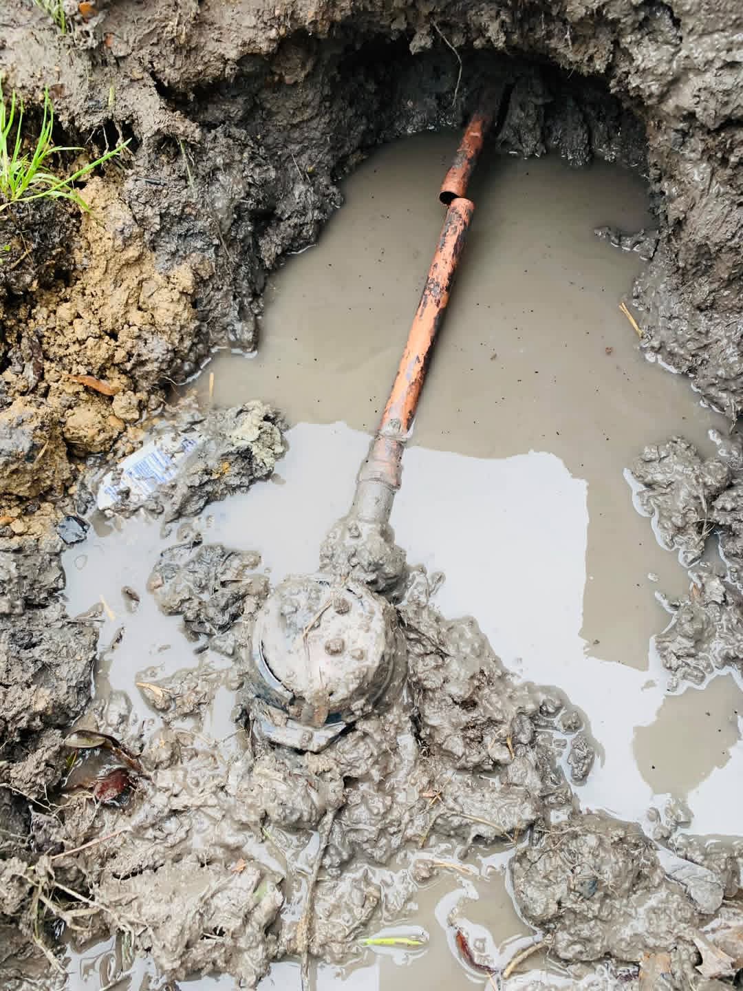 A rusty pipe connected to a valve lies in muddy water within a hole in the ground.