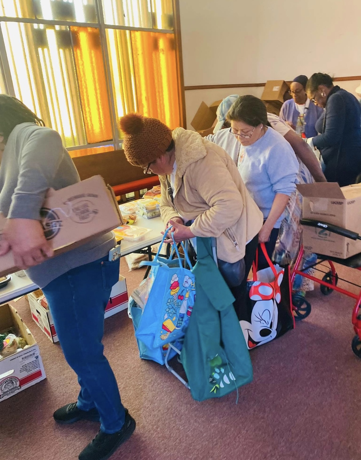 People standing in line at a table with boxes, bags, and items, near a window with orange and yellow stained glass.