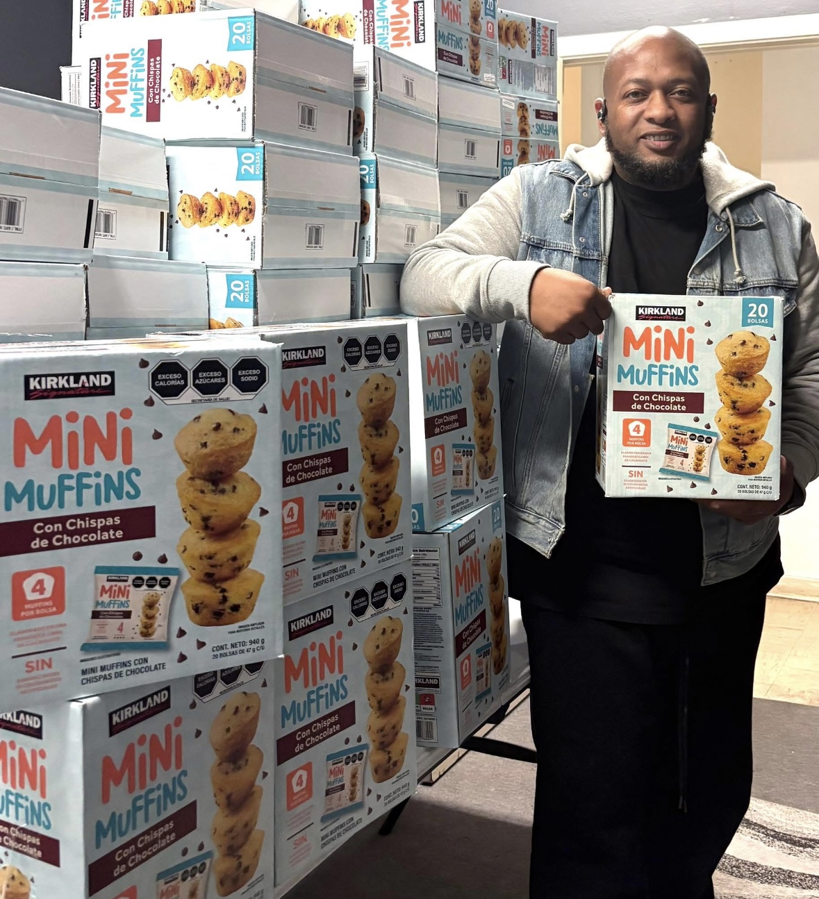 Man holding a box of mini muffins in a store aisle with stacked muffin boxes around him.