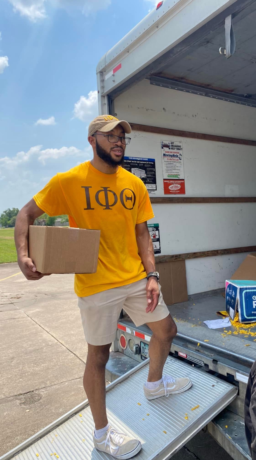 Person wearing yellow shirt with Greek letters, beige shorts, white sneakers, and a cap, standing outside near a truck.