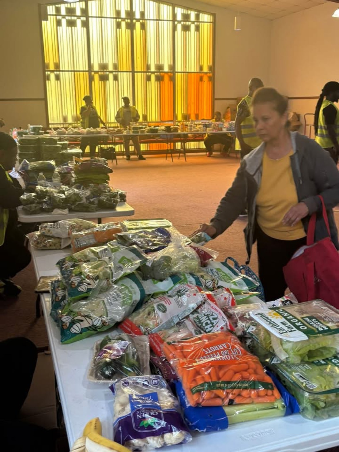 Woman shopping at a table with packaged vegetables and food items in a large indoor space with colorful window panels.