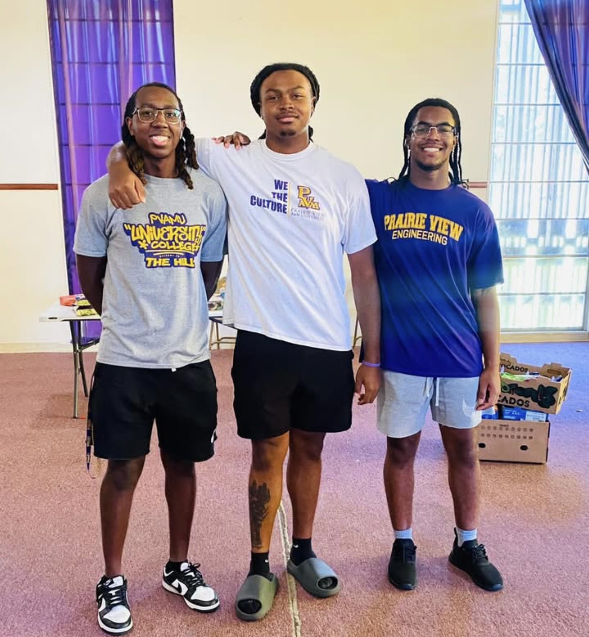 Three young men standing indoors, arms around each other, smiling, with purple curtains and windows in the background.